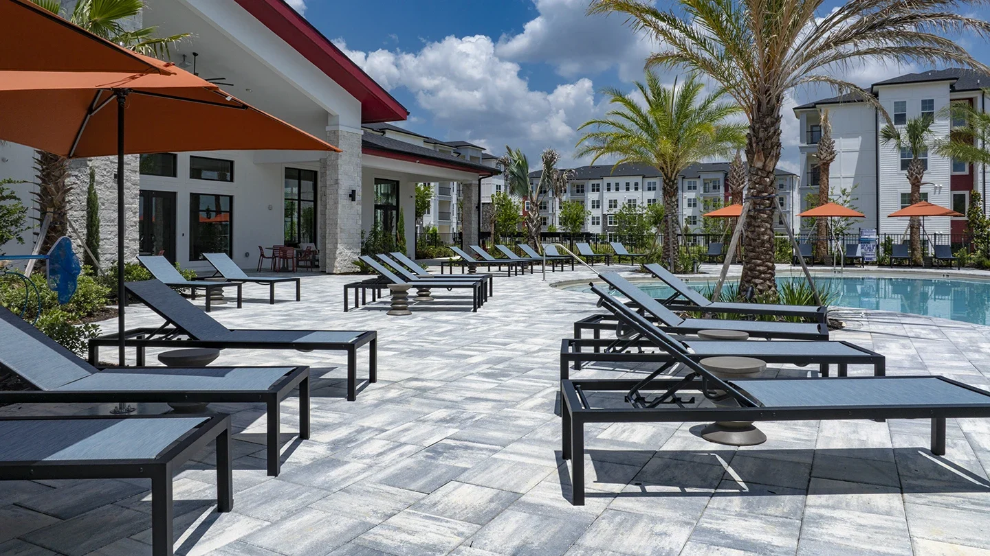 Spacious pool deck featuring rows of modern black-framed lounge chairs with mesh fabric and small round side tables, bordered by palm trees, orange umbrellas, and apartment buildings around a clear blue swimming pool.