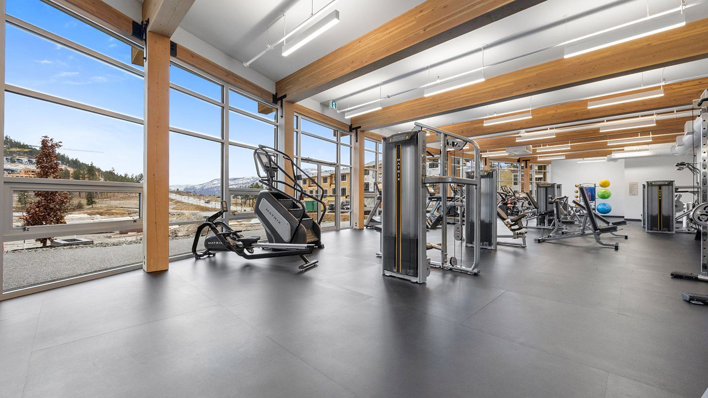 A bright, modern fitness centre with high ceilings and large floor-to-ceiling windows offering a scenic view of rolling hills and residential buildings. The room features exposed wood beams, LED lighting, and black rubber flooring. Various gym equipment is neatly arranged, including a stair-climber, weight machines, stationary bikes, and a set of colorful stability balls mounted on the back wall.