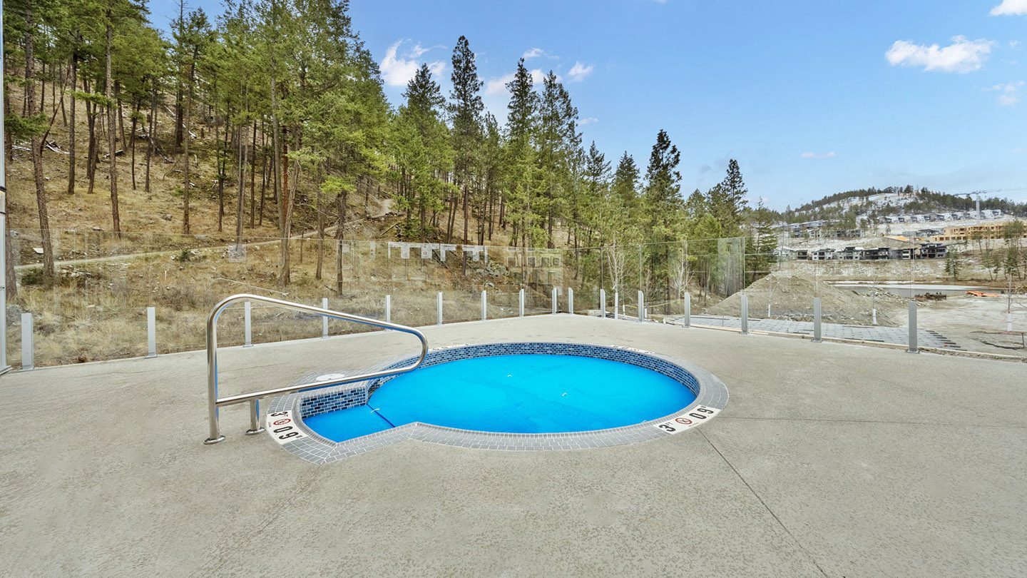 A circular outdoor hot tub surrounded by a textured concrete deck, enclosed by a clear glass safety fence. The hot tub is equipped with a stainless steel handrail and marked depth indicators ranging from 3 to 6 feet. The area is set against a backdrop of a wooded hillside with tall pine trees, and there are distant views of ongoing residential development and forested hills under a clear blue sky.
