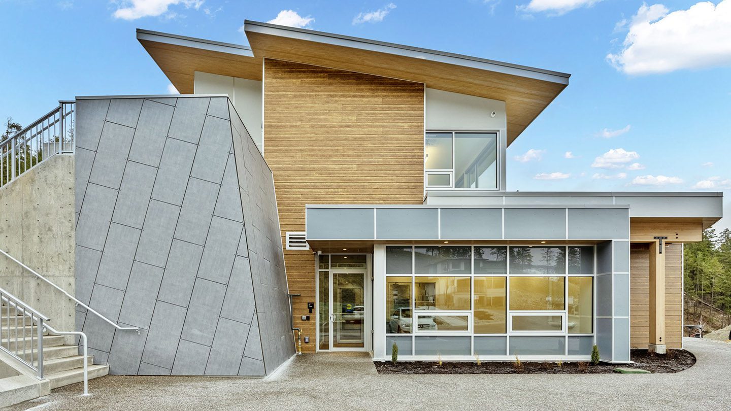 Modern two-storey amenity building featuring a mix of natural wood siding and grey architectural panels. The building has large windows, a glass entrance door, and an angled roofline. To the left, a concrete staircase with metal railings leads to the upper level. The surrounding area includes a gravel pathway, landscaped beds, and forested hills under a partly cloudy blue sky.