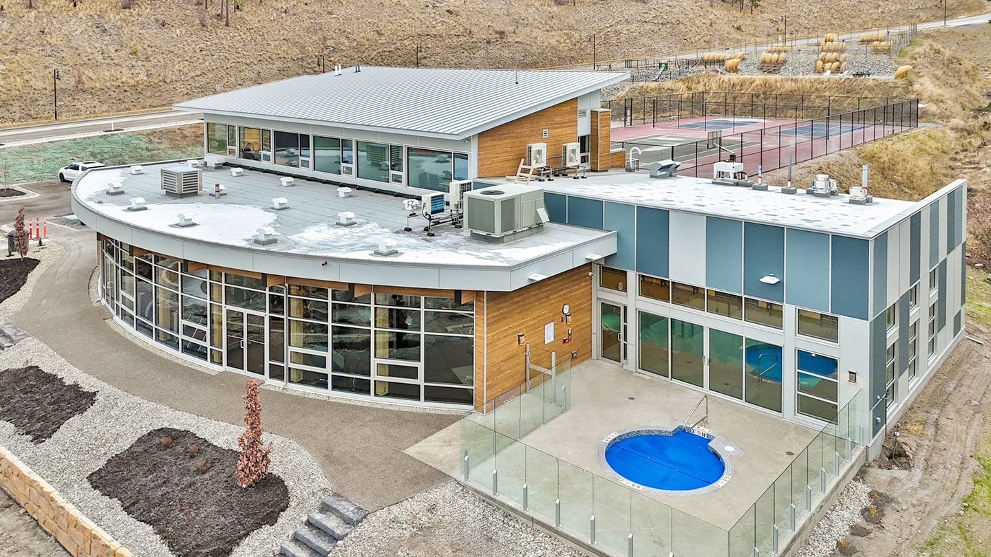 A modern recreational facility with a mix of wood and glass exterior, including large floor-to-ceiling windows and a curved roof section. In the foreground, a circular outdoor hot tub is enclosed by a glass fence. Indoor pool facilities are visible through the glass doors. Behind the building, fenced tennis and pickleball courts are located on a terraced hillside landscaped with gravel and ornamental grasses.