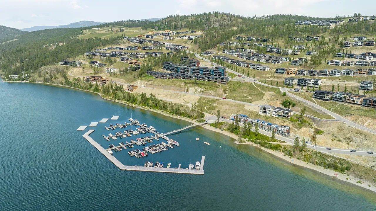Aerial view of McKinley Beach on the shore of a large lake, showing a modern marina with multiple boat slips extending into the water. Behind the marina, terraced hills are lined with contemporary homes and multi-level residential buildings surrounded by pine forest. Curving roads wind through the development, connecting the waterfront to higher-elevation residences.