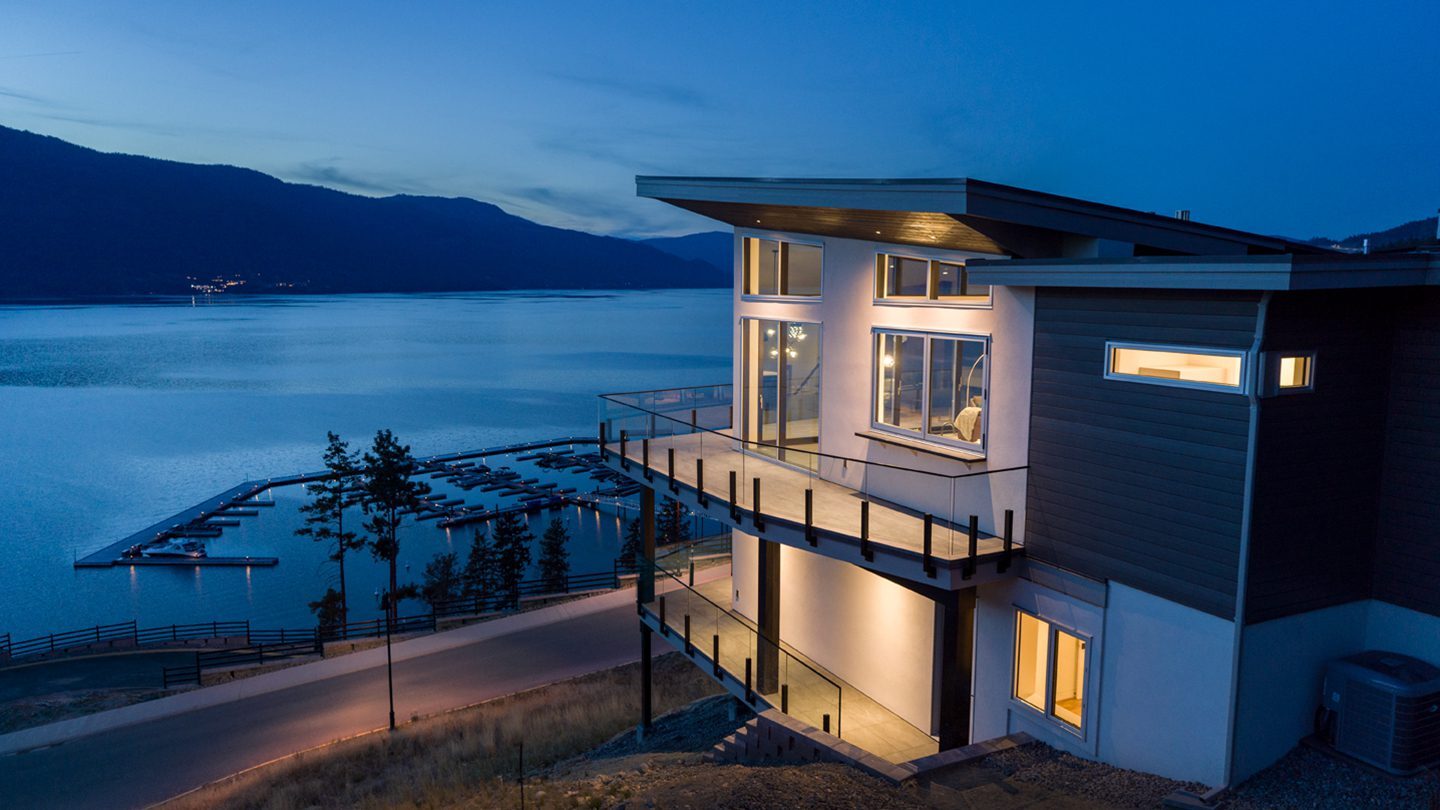 A modern, multi-level home with large windows and wraparound balconies overlooks a serene lake at dusk. The house features a flat roof, a mix of white and dark grey exterior panels, and glass railings. Below, a marina with several docks extends into the calm blue water. Soft lighting glows from the home