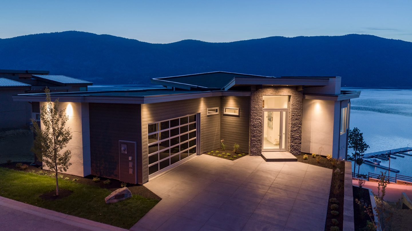 A contemporary single-story home with a flat roof and large glass garage door is softly lit at twilight. The house features dark horizontal siding, stone accent walls, and a well-manicured front lawn. The home overlooks a calm lake with a marina below, and forested mountains rise in the distance under a dusky blue sky.