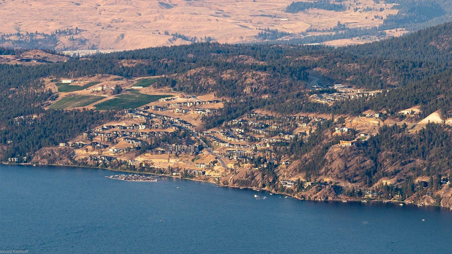 Aerial view of the McKinley Beach residential development nestled in the hills above a large blue lake. The community features clusters of modern homes and buildings built along winding roads and terraced slopes. Surrounding the development are densely forested areas, with a patchwork of green fields and dry, rugged terrain in the distance.