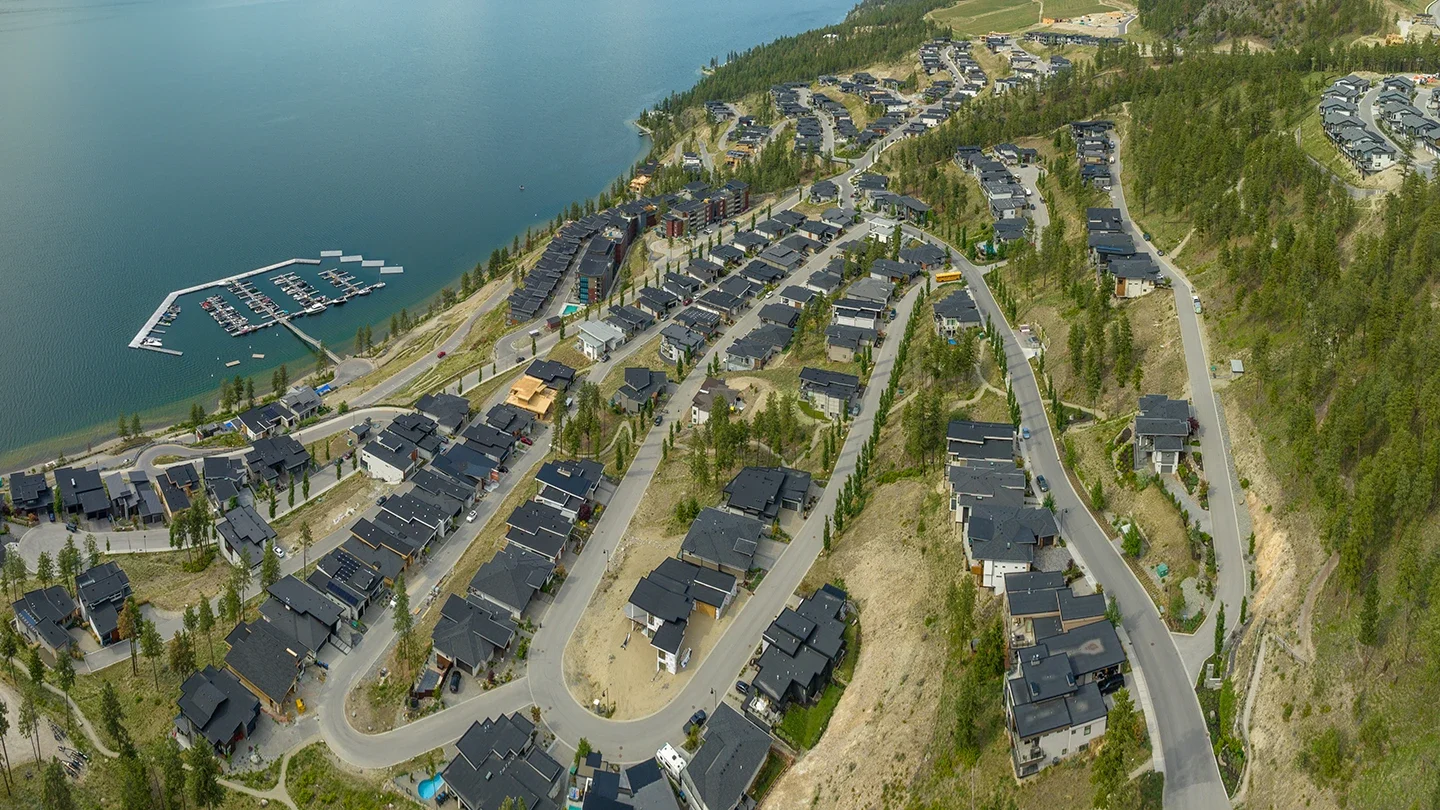 Aerial view of McKinley Beach featuring curving streets lined with modern homes and townhouses. The development is built into hilly terrain with sparse tree cover, and a large marina with numerous boats extends into the lake. The image captures a mix of completed homes and ongoing construction, surrounded by forested areas and rising hills in the background.