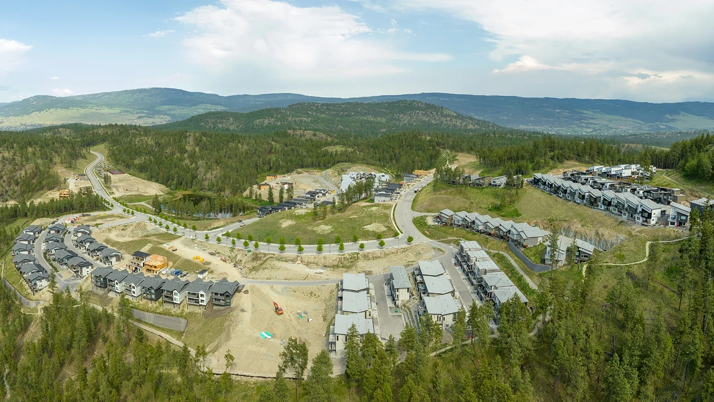 A panoramic aerial view of McKinley Beach surrounded by forested land and mountains. The image shows multiple clusters of modern townhomes and detached houses in various stages of construction. A winding road runs through the middle of the community, with a small pond and patches of green space visible. In the background, tree-covered hills stretch to the horizon under a partly cloudy sky.