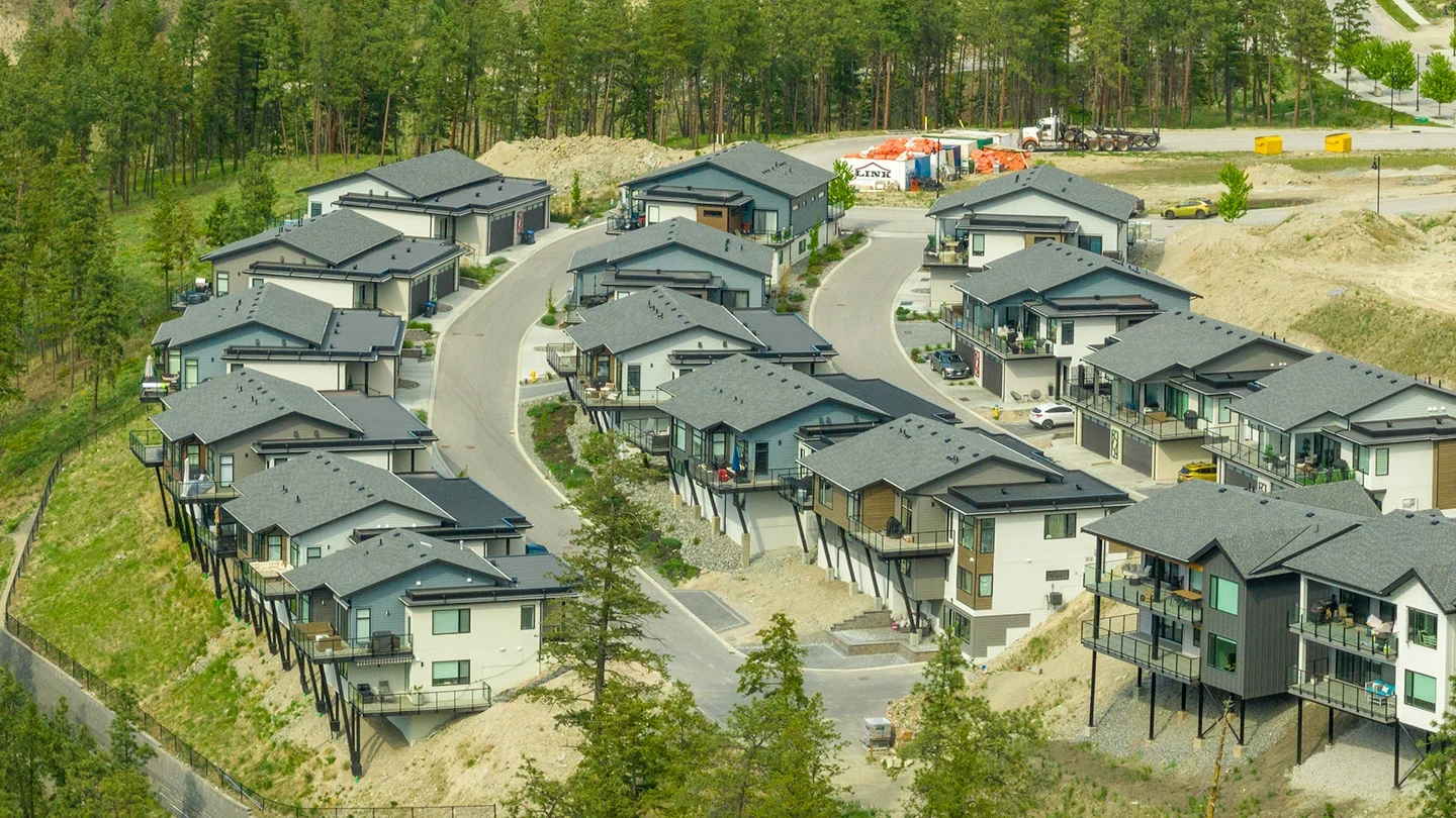 A close-up aerial view of McKinley Beach featuring rows of two-story homes built on steep terrain. Many houses are supported by tall stilts to accommodate the sloped hillside. The street curves gently through the area, and driveways and garages are visible. Behind the homes is a lightly forested area and construction staging with visible materials and equipment.