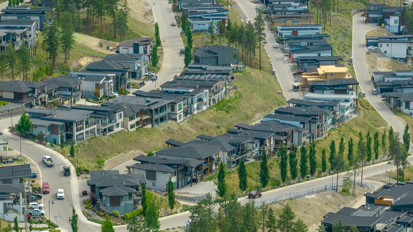 An aerial view of McKinley Beach showing a series of modern, flat-roofed homes built along curved roads on a hillside. The homes are arranged in parallel rows with manicured landscaping, driveways, and some homes under construction. A line of young trees follows a road in the foreground, and forested land surrounds the area.