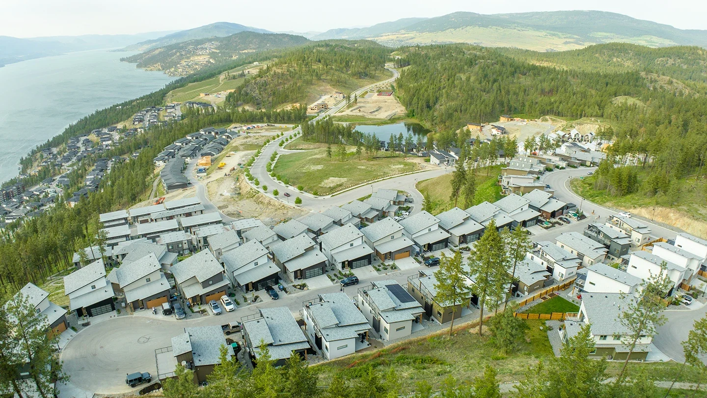 An expansive aerial view of McKinley Beach overlooking Okanagan Lake, with rows of modern grey-roofed homes set along curving roads. The lake is visible on the left side, bordered by forested terrain. A pond and additional homes are situated in the central background, surrounded by rolling green hills and forested mountains under a lightly clouded sky.