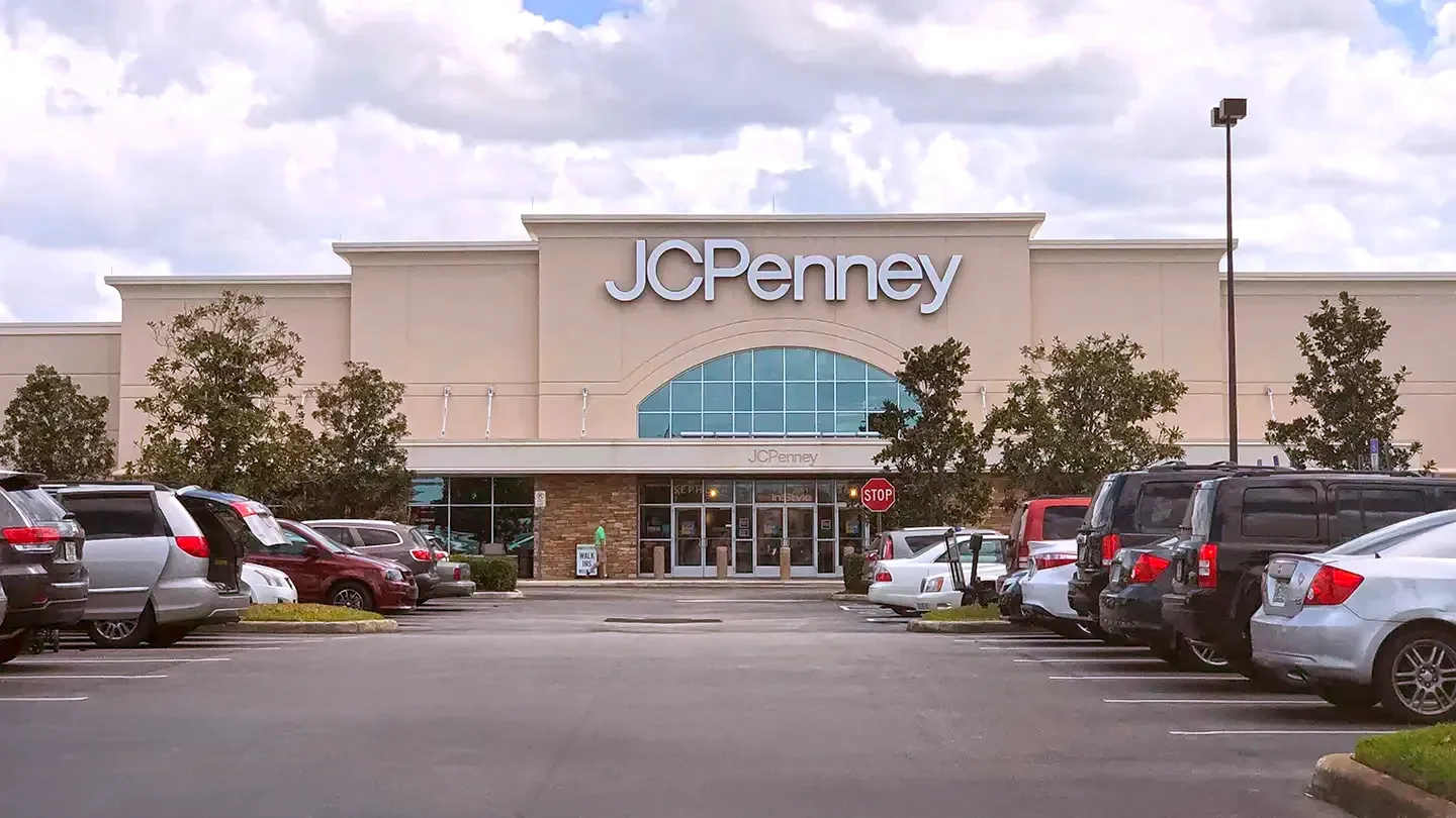 Street-level view of a JCPenney storefront with large white lettering above a central glass archway. The building has a light beige facade with brickwork around the entrance. Multiple trees are planted along the front of the store. A busy parking lot is in the foreground, with several parked cars and a person walking toward the entrance. A red stop sign and a "Walk-Ins Welcome" sign are also visible near the store’s front doors. The sky is partly cloudy.