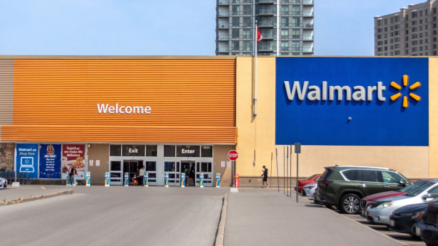 Front view of a Walmart entrance with large blue and orange signage, glass sliding doors labeled “Exit” and “Enter,” and a row of parked vehicles in front.
