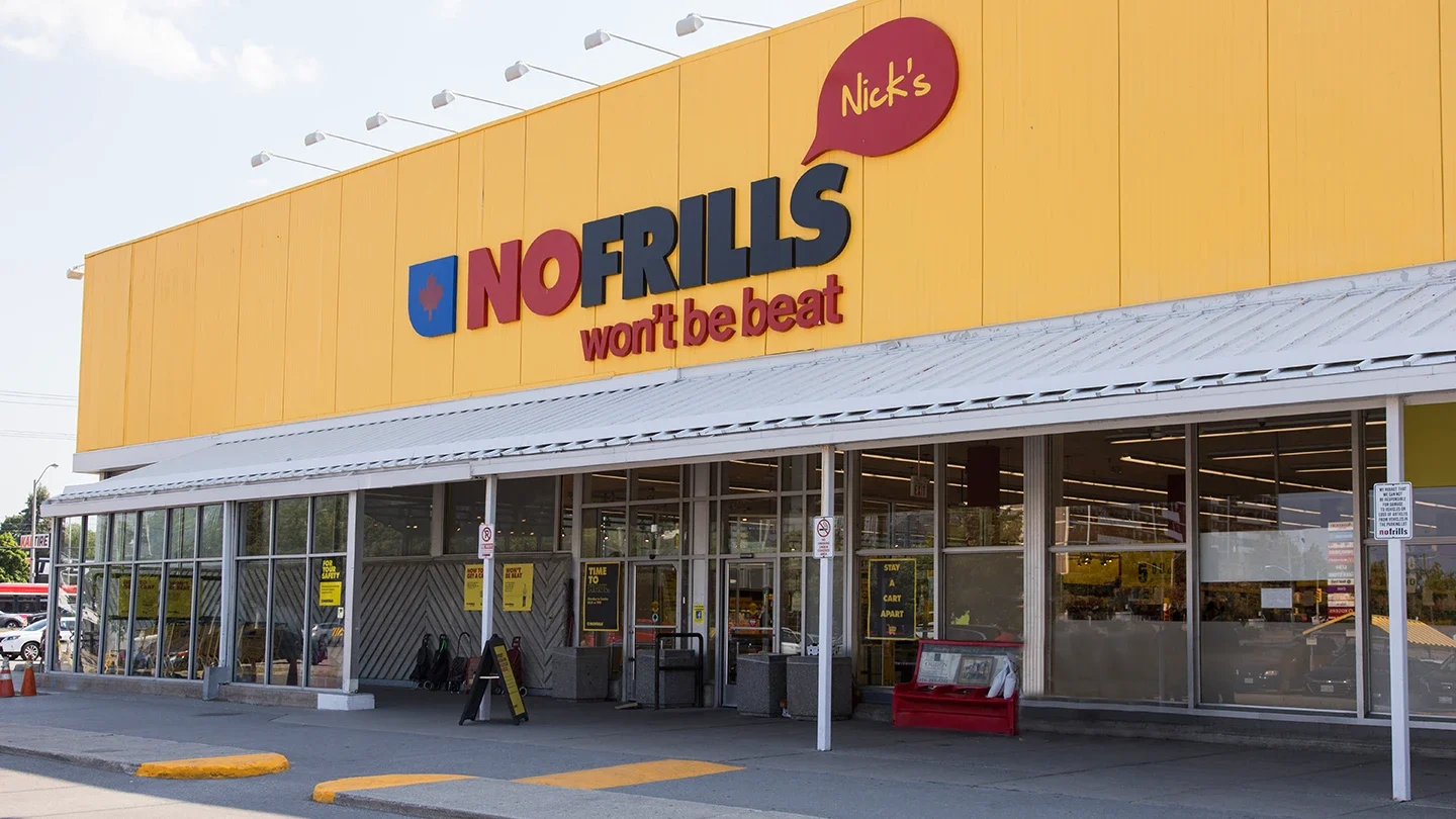 Exterior view of Nick’s No Frills with a bright yellow facade, red and blue branding, and large glass windows showing grocery store interior.