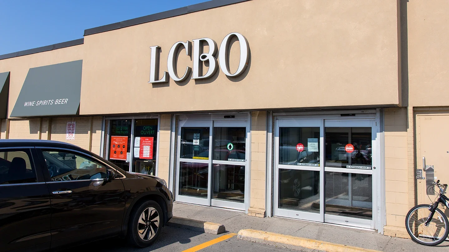 Street-level image of an LCBO location with beige brick walls, green awnings labeled “Wine • Spirits • Beer,” and a sliding glass double-door entrance.
