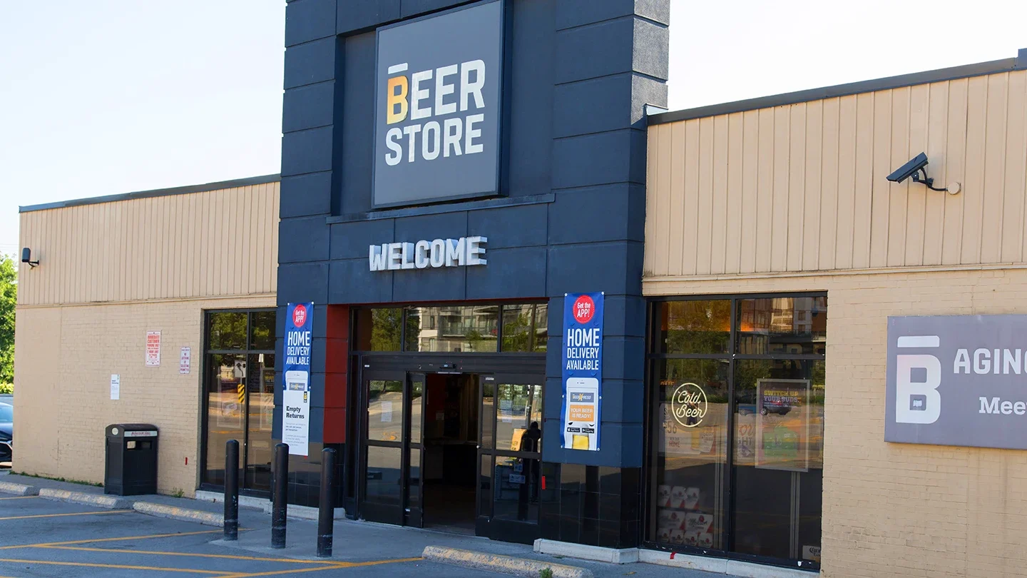 Street-level view of a Beer Store entrance with a prominent “Welcome” sign above double glass doors, home delivery signage, and light tan brick walls.