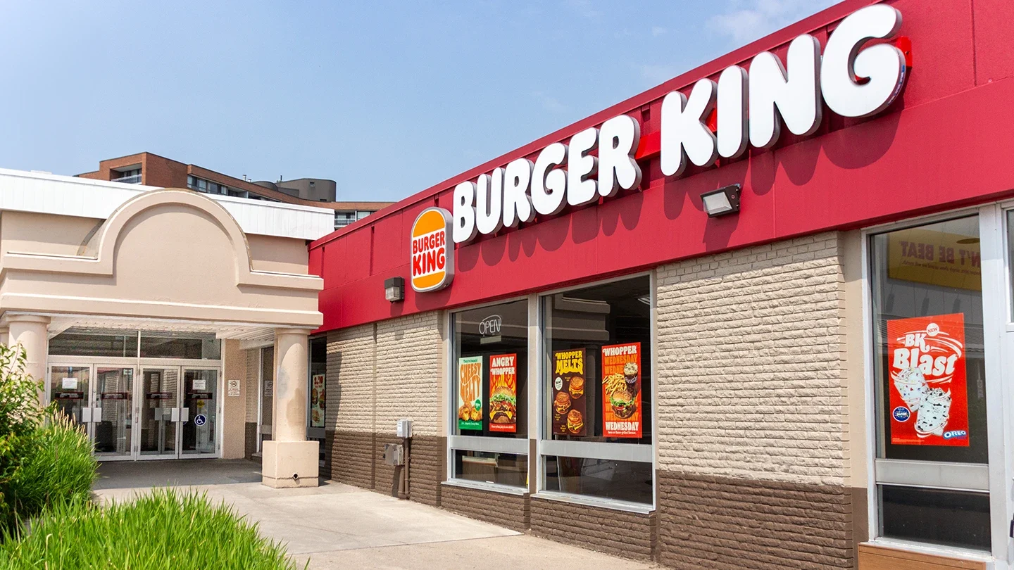 Close-up view of a Burger King storefront with red trim and large white letters above windows showcasing posters of menu items, next to an arched entrance to the mall.