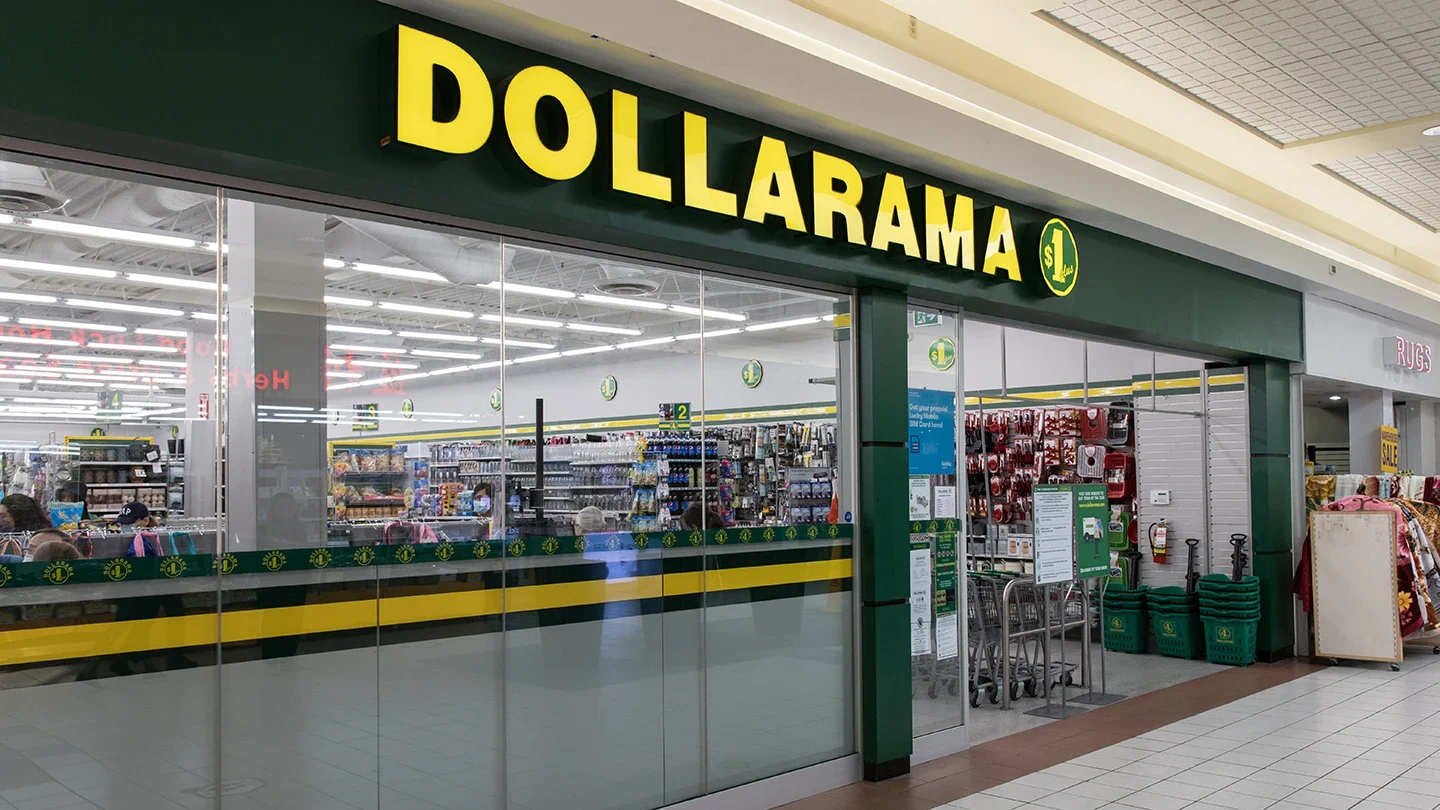 Interior view of a Dollarama store in a shopping mall, featuring a wide entrance with glass panels and visible shelving stocked with products inside.