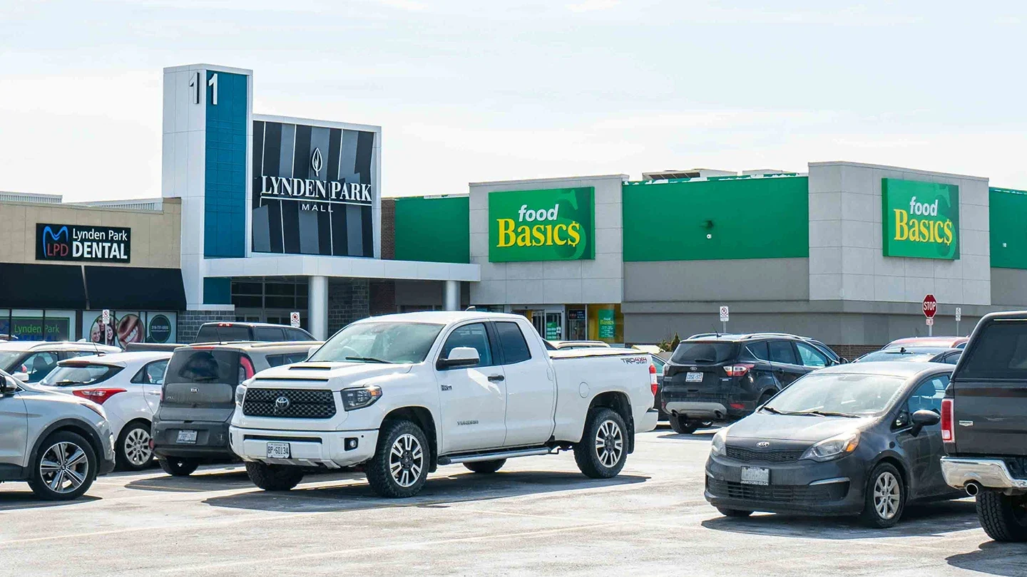 Front view of Lynden Park Mall showing the main entrance with a sign labeled "Lynden Park Mall" and adjacent storefronts for Food Basics and Lynden Park Dental. Several cars are parked in the lot under a clear sky.