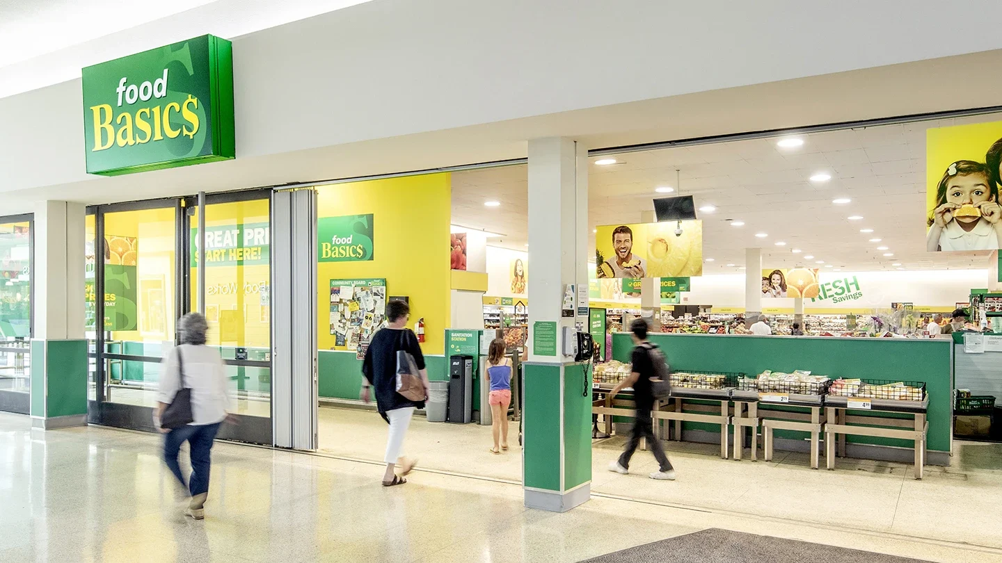 Front entrance of a Food Basics grocery store located inside a shopping mall. Several people are walking in and out of the store. The interior displays fresh produce and promotional signage with bright yellow and green branding.