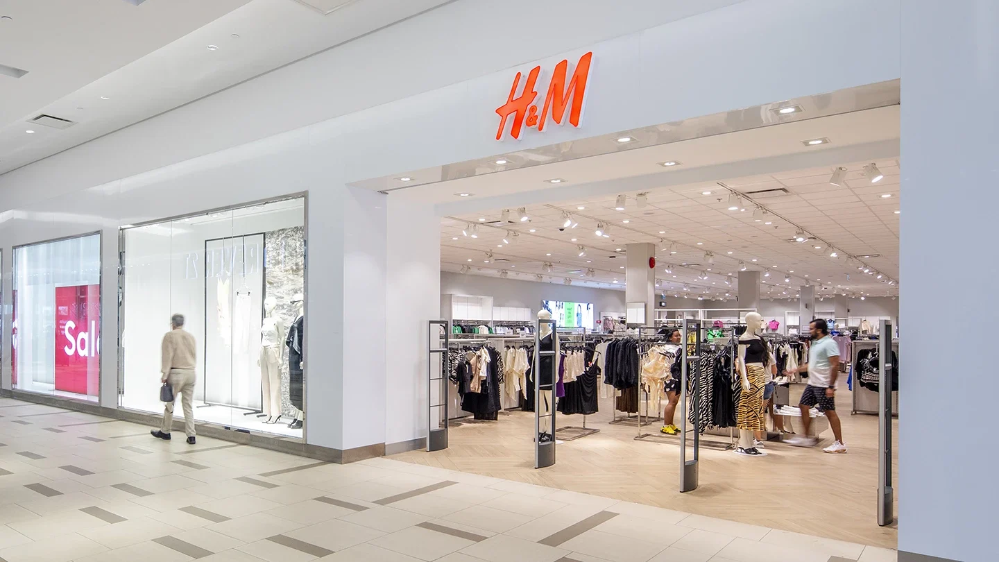 Entrance to an H&M clothing store inside a mall. Shoppers walk past mannequins and clothing racks visible through the wide open storefront. Bright lights and organized displays create a modern retail atmosphere.