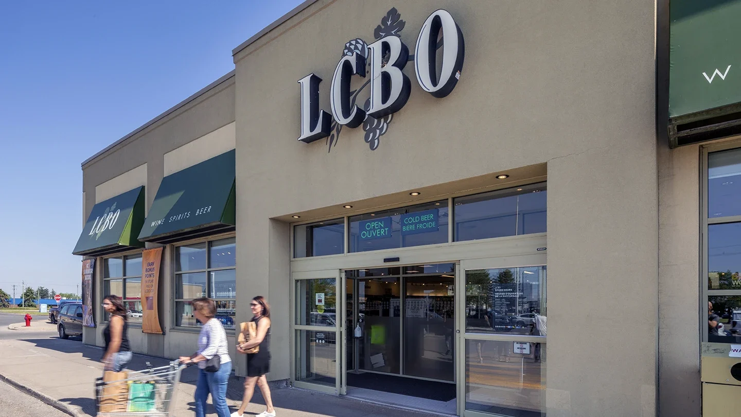 Exterior of the LCBO store at Lynden Park Mall, with green awnings and signage reading "Wine Spirits Beer", as shoppers with carts and bags walk past the entrance.