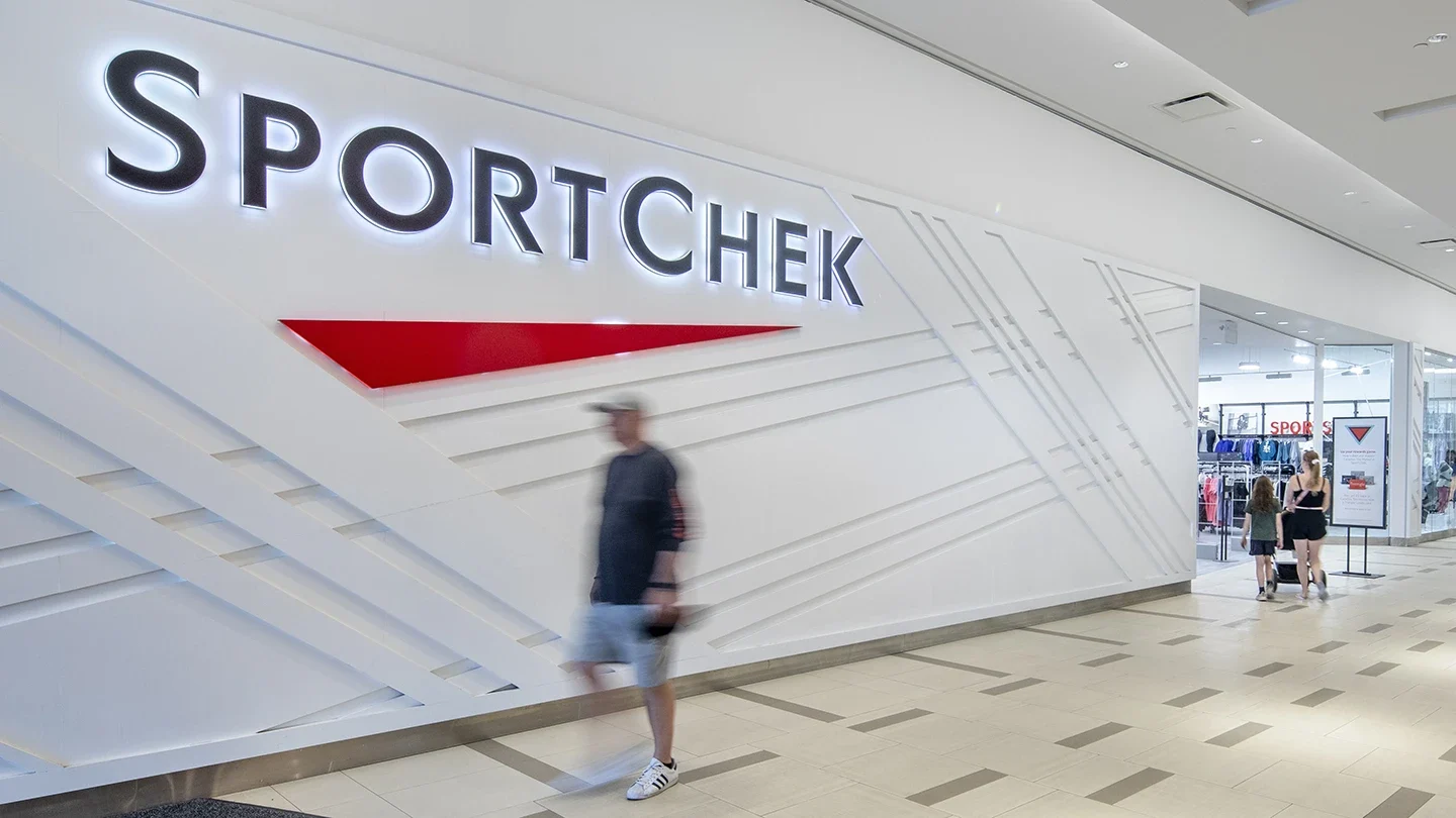 Interior mall view of Sport Chek storefront with large modern white and red branding on the wall and shoppers walking past the entrance.