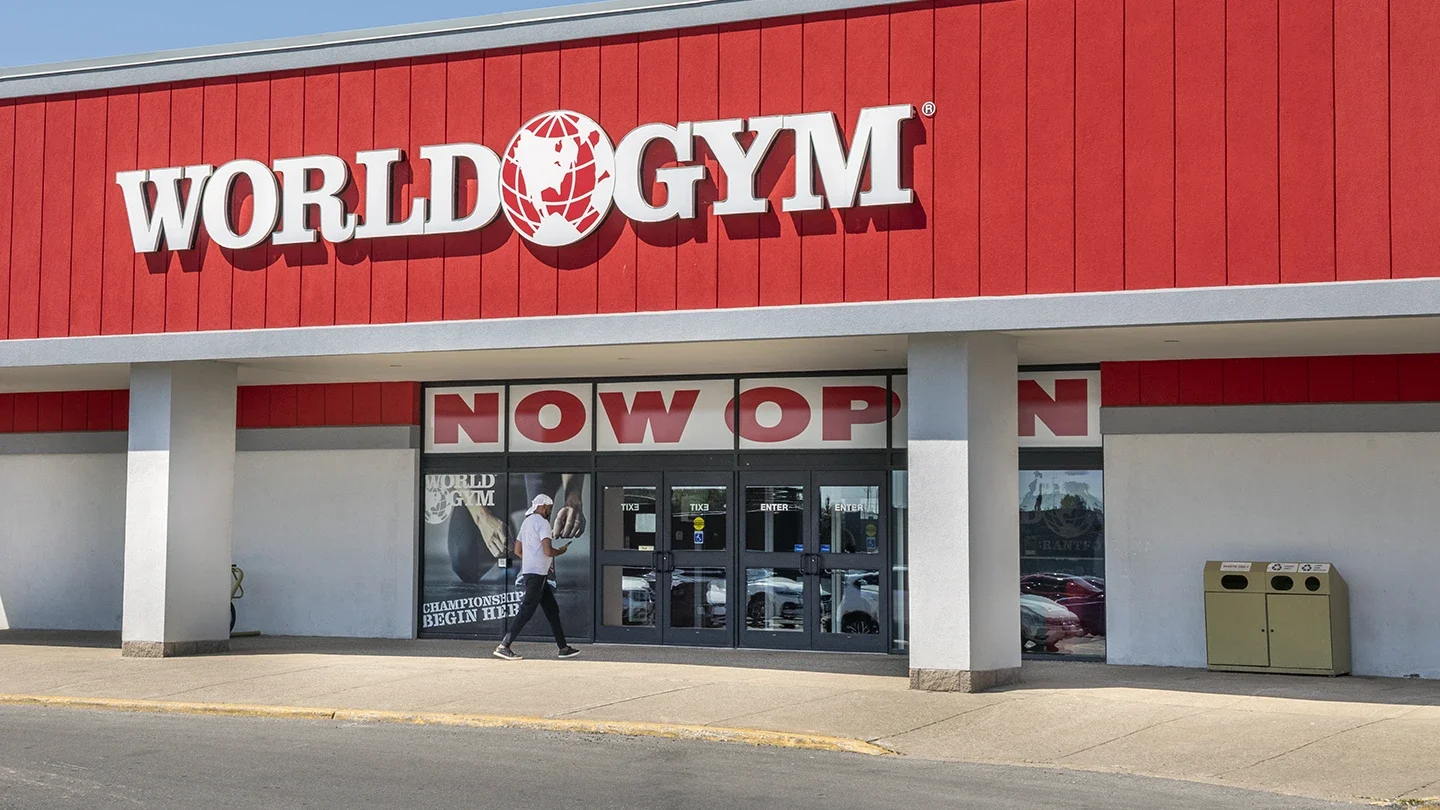 Exterior view of World Gym with red facade and large "Now Open" signage across the windows, and a person walking past the entrance.