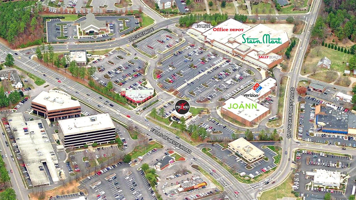 A labeled aerial image of The Falls Center retail plaza, showing surrounding roads and parking lots. Notable businesses are marked, including Office Depot, Stein Mart, Sally Beauty, Verizon, Joann, Domino’s, Pei Wei Asian Diner, and Anytime Fitness. Major roads like Falls of Neuse Road and Old Wake Forest Road are identified.