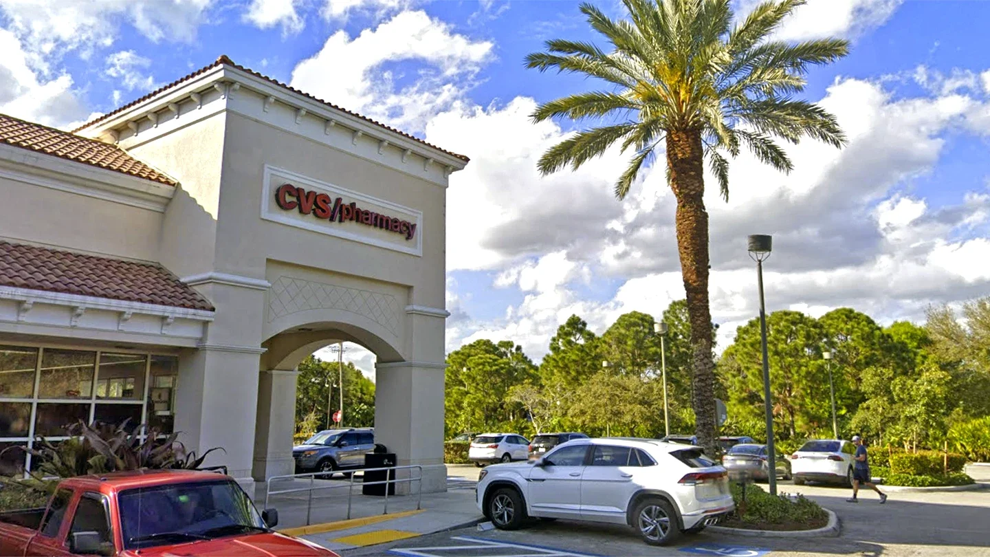 Ground-level image of a CVS/pharmacy storefront with a red pickup truck and white SUV parked in front, a tall palm tree to the right, and a partly cloudy sky with trees in the background.