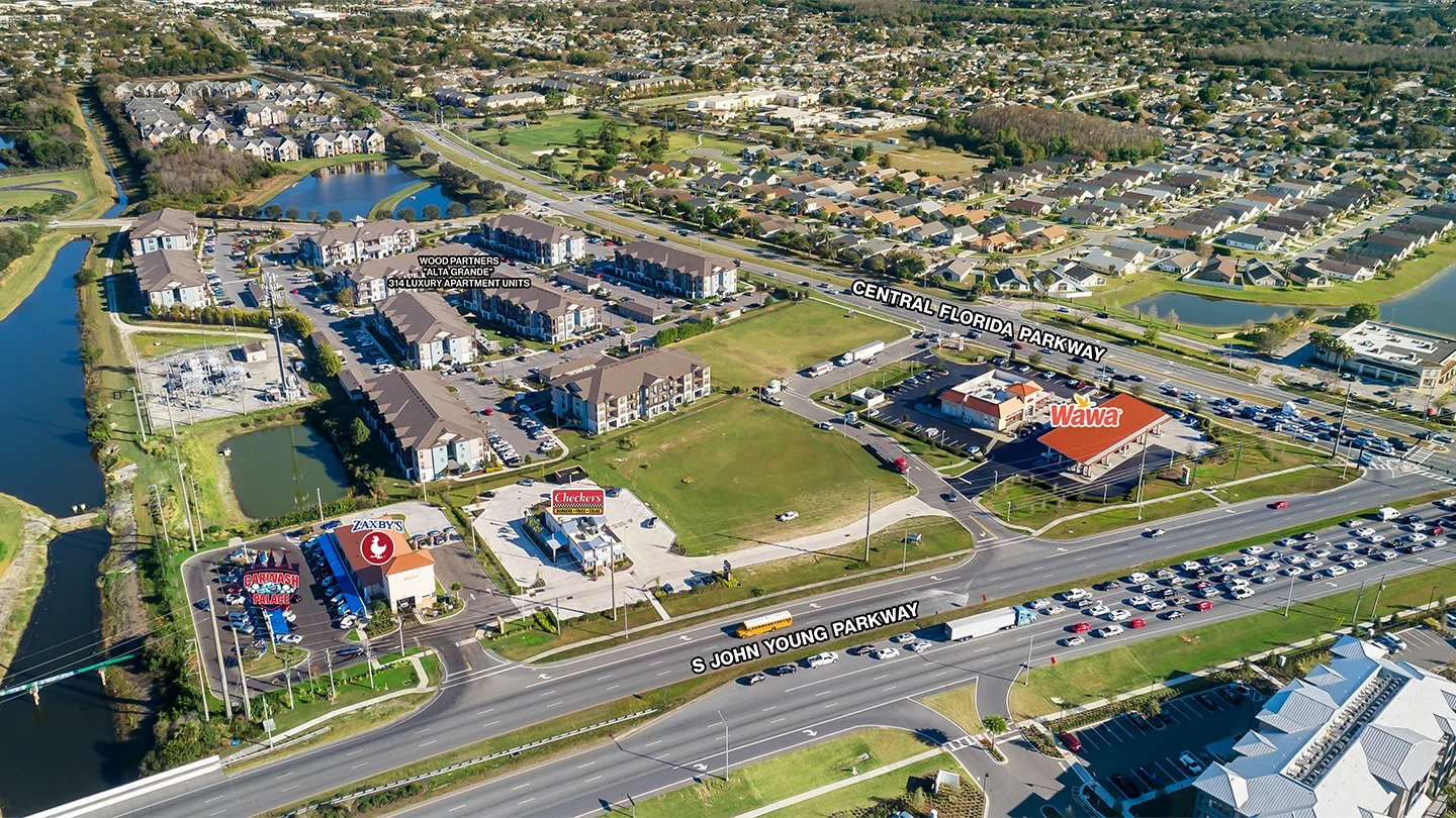 A labeled aerial view of Grande Lakes Plaza showing the Wawa building and nearby retailers such as Zaxby’s, Checkers, and a car wash, surrounded by apartment complexes and roadways including Central Florida Parkway and S John Young Parkway.
