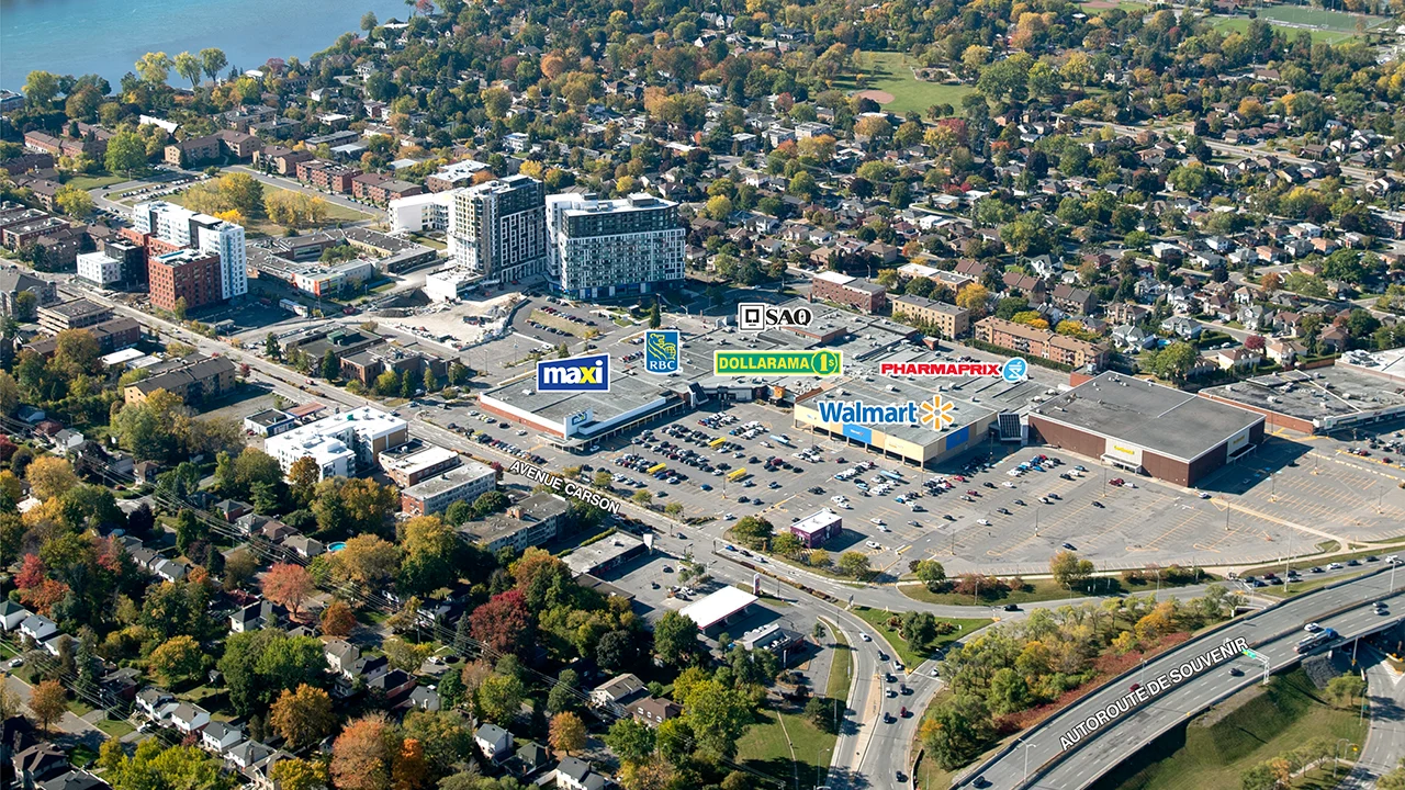 Aerial view of the Les Jardins Dorval shopping centre in Dorval, Quebec, showing a large commercial complex with several major retail stores and a spacious parking lot. Storefronts labeled include Walmart, Maxi, Dollarama, Pharmaprix, RBC, and SAQ. The complex is bordered by Avenue Carson to the south and Autoroute de Souvenir running across the bottom of the image, with several vehicles visible on both roads. A high-rise residential or office building and a construction crane are visible behind the shopping centre. The surrounding area includes residential neighborhoods with trees and low-rise buildings, parks, and visible river waterfront in the upper part of the image.