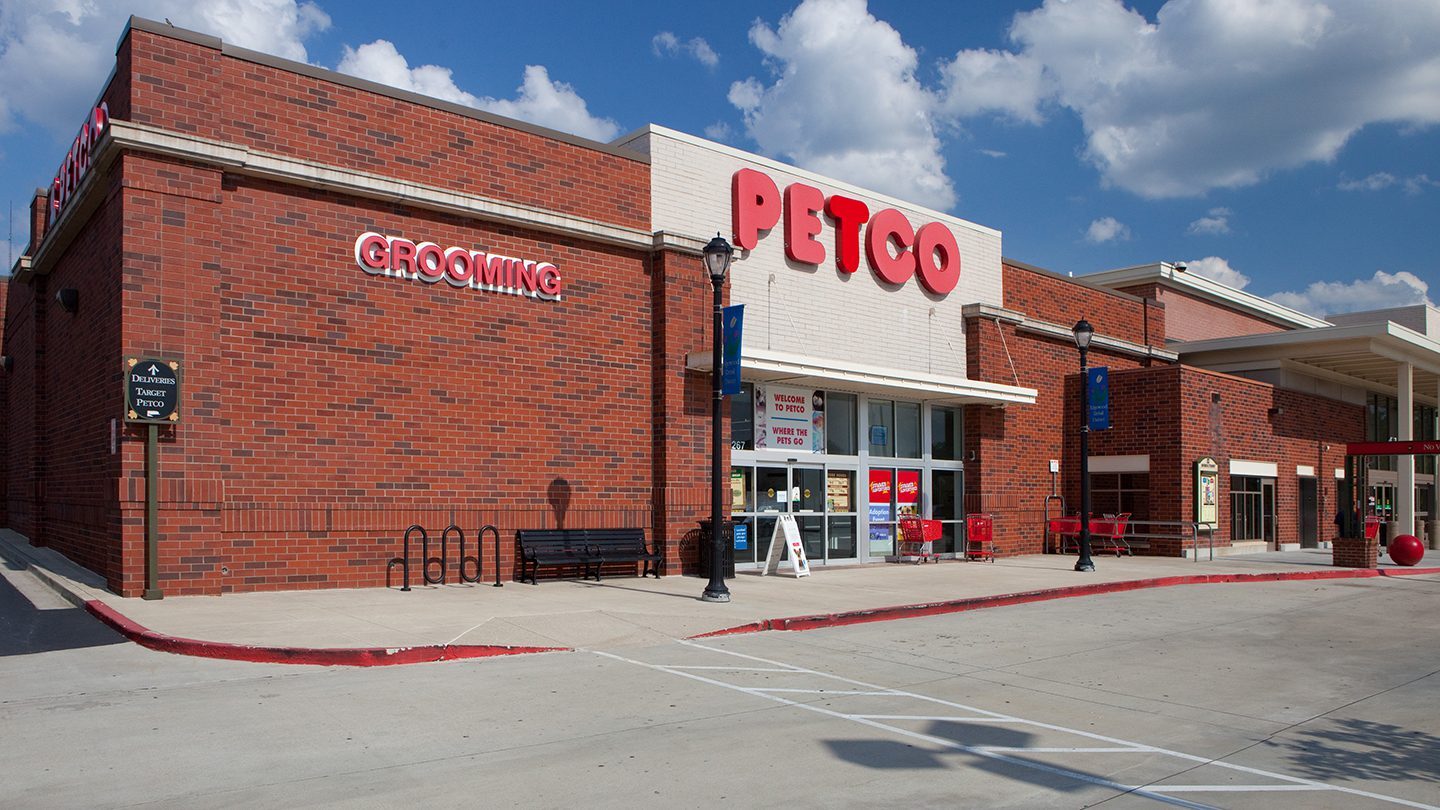 Daytime view of the Petco store exterior at the Edgewood Retail District in Atlanta, Georgia. The building features red brick with a white accent facade displaying the large red “PETCO” logo above the entrance. The word “GROOMING” is mounted on the side of the building. Red shopping carts are lined up near the doors. A blue sky with scattered white clouds is visible above, and there are lamp posts and a bike rack near the sidewalk in front of the store.