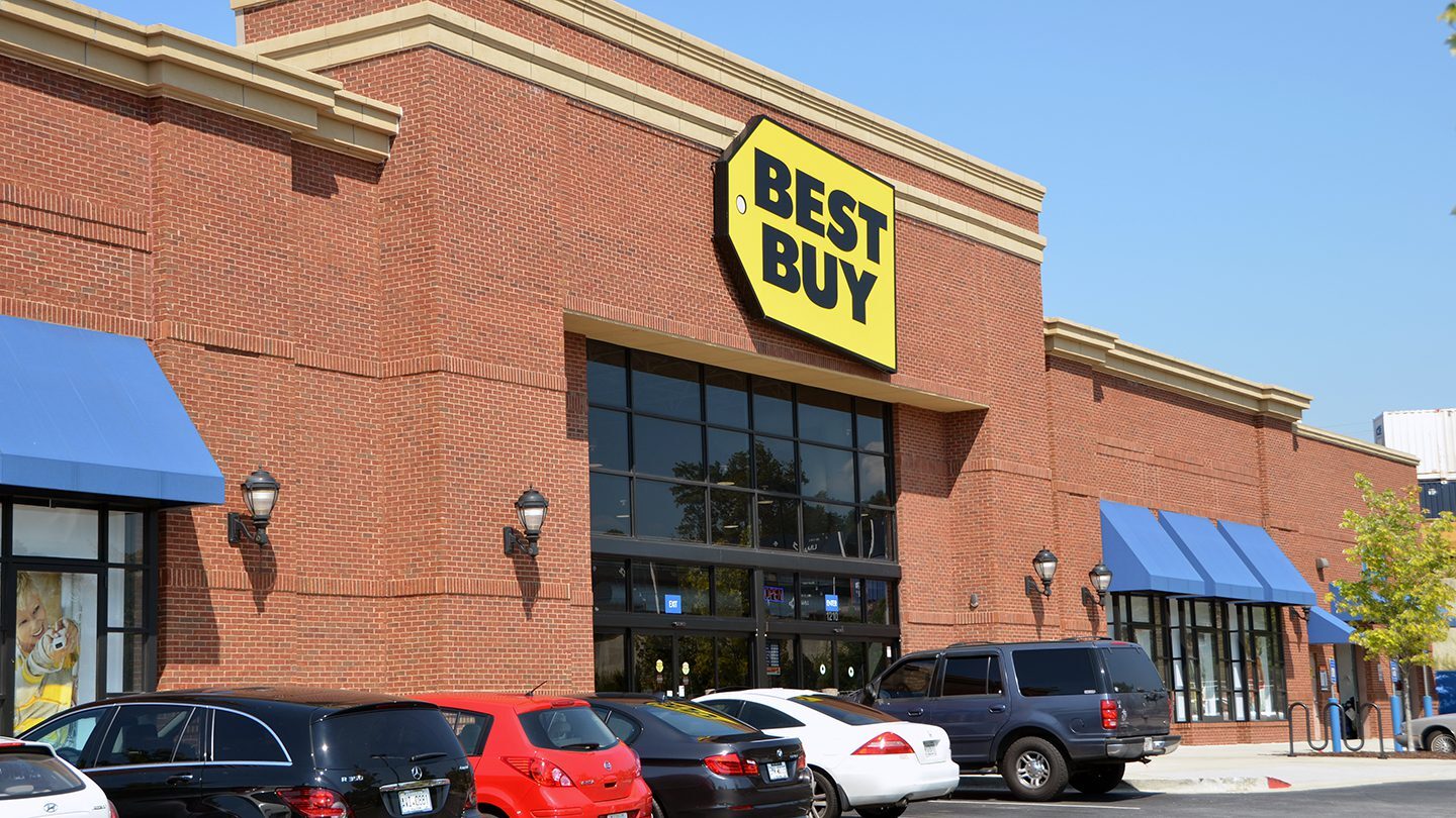 Exterior view of the Best Buy store located in the Edgewood Retail District in Atlanta, Georgia. The building features a prominent yellow and black Best Buy sign mounted above large, black-framed windows and double entrance doors. The facade is constructed of red brick with decorative beige trim. Several blue awnings cover adjacent windows, and multiple cars are parked in front of the entrance under a clear blue sky.