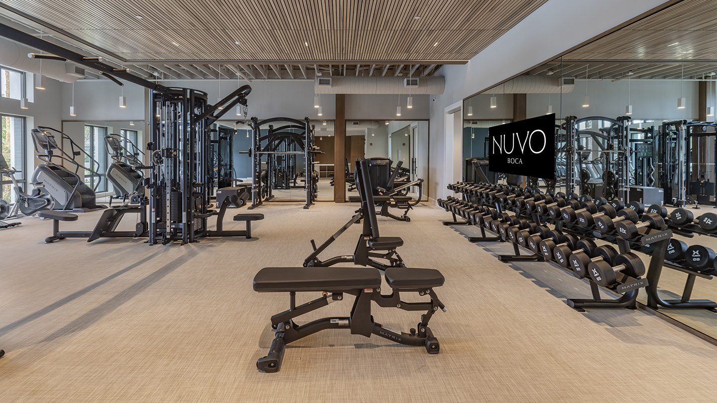Spacious and modern gym with a variety of exercise equipment, including treadmills, elliptical machines, weight benches, free weights, and multi-use strength training machines. The space features floor-to-ceiling mirrors, a wooden slat ceiling, and the “NUVO Boca” logo displayed on a mirrored wall.