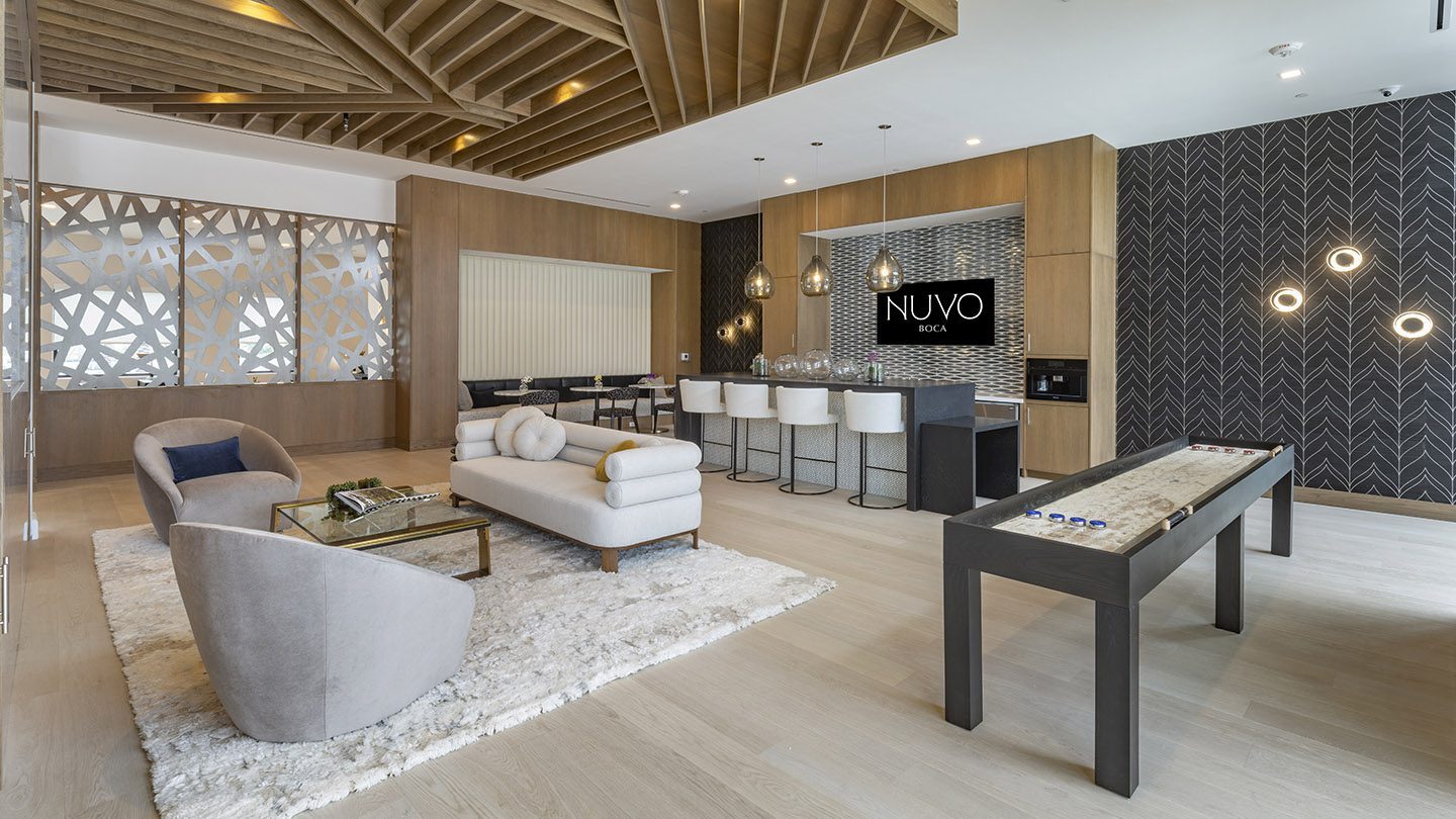 Elegant lounge with four blue velvet armchairs arranged around a coffee table on a black-and-white checkered tile area. To the right, a tall built-in bookshelf features decorative books, plants, and artistic objects with warm accent lighting. Large glass doors and windows provide a view of the outdoor pool area and palm trees.