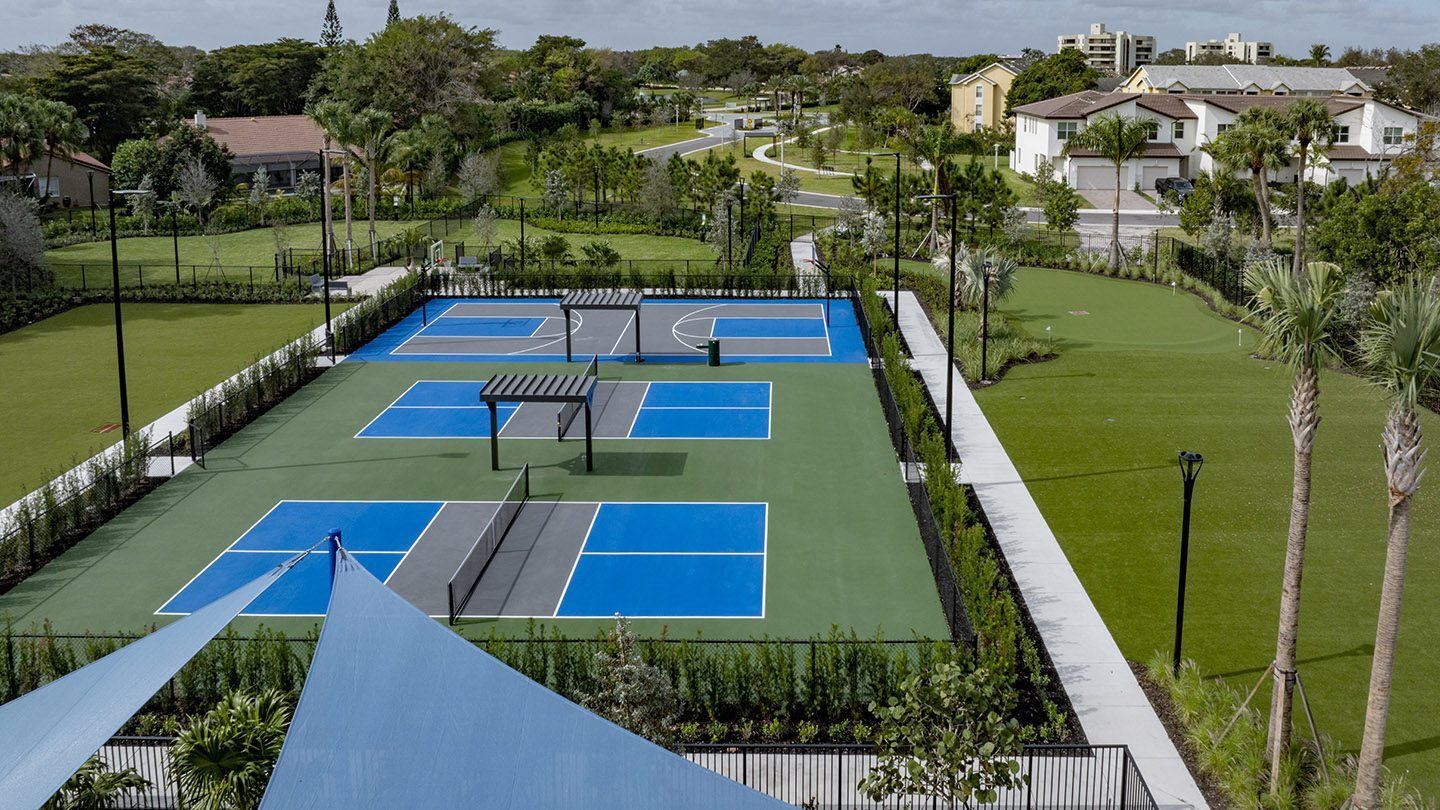 Aerial view of outdoor recreational area featuring vibrant blue and green pickleball courts, a basketball court, and shaded seating areas. Adjacent to the courts is a well-maintained putting green surrounded by palm trees and walking paths. The entire facility is enclosed by fencing and lush landscaping, with residential homes and a winding road visible in the background.
