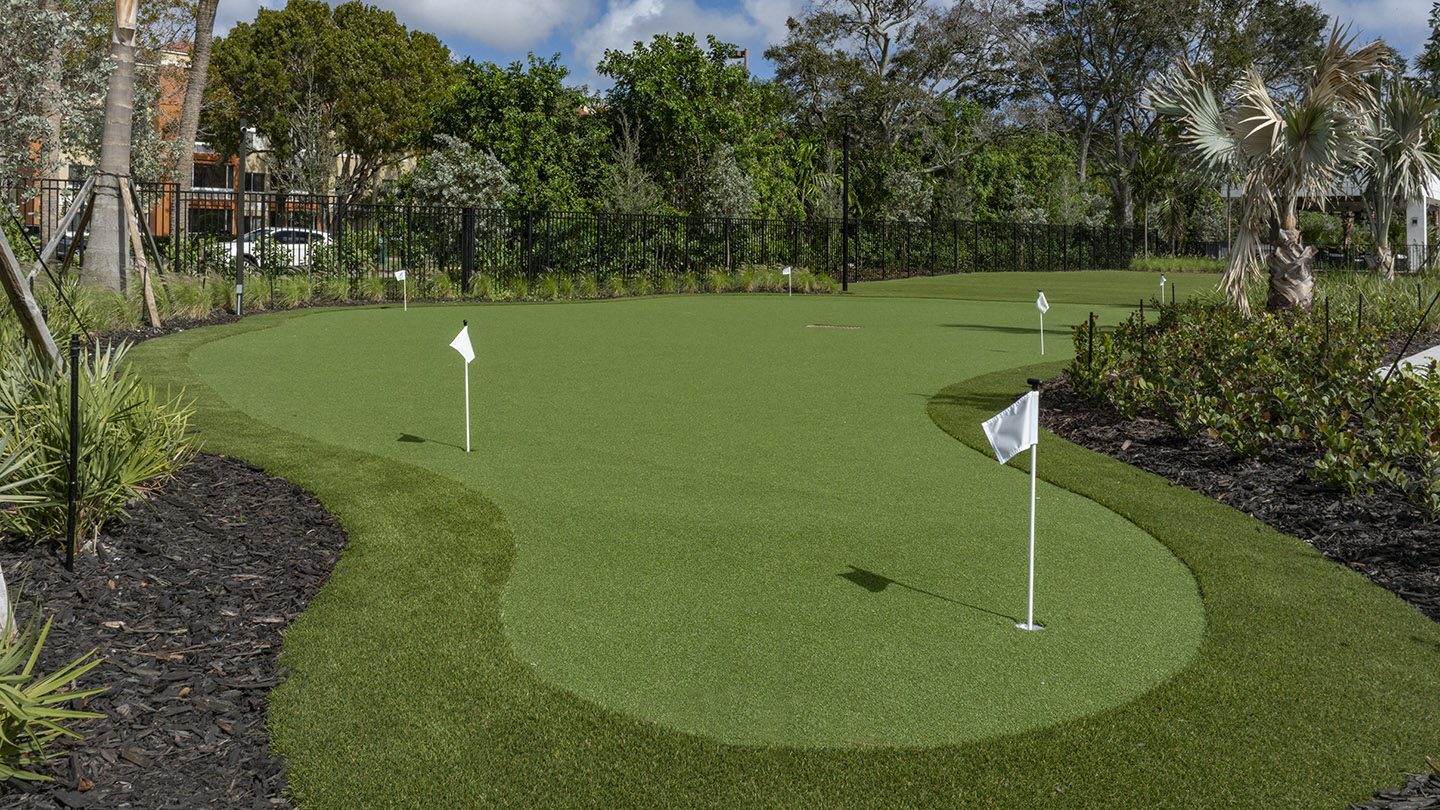 Close-up view of a professionally manicured putting green featuring multiple holes marked by white flags. The green is bordered by landscaped mulch beds, tropical plants, and palm trees. A black metal fence and lush trees provide a serene, natural backdrop under a bright blue sky with scattered clouds.