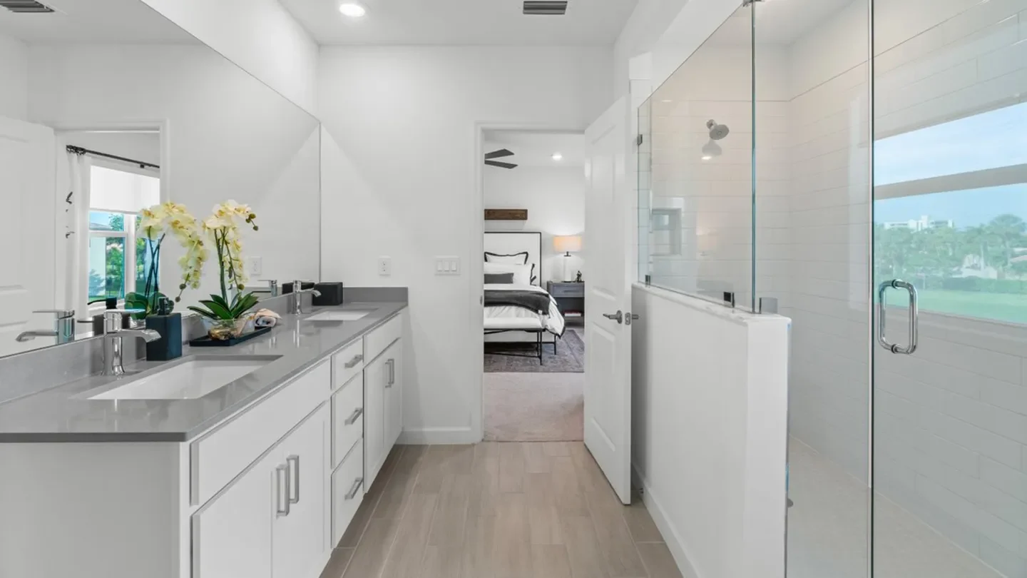 Bright, contemporary bathroom featuring a double-sink vanity with a gray countertop, white cabinetry, and chrome fixtures. A large mirror spans the wall above the sinks, and decorative orchids add a natural touch. On the right side, a spacious glass-enclosed walk-in shower is visible. The room leads into a bedroom with a neatly made bed and bedside lamp, seen through an open doorway.