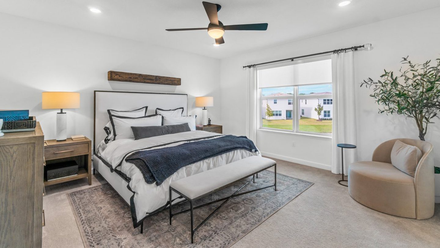Bright, modern bedroom featuring a large bed with white and navy bedding, two matching bedside tables with lamps, and a soft area rug. A cushioned bench sits at the foot of the bed. To the right, a large window with white curtains allows natural light to fill the space. A round beige armchair, small side table, and decorative plant enhance the cozy and stylish atmosphere.