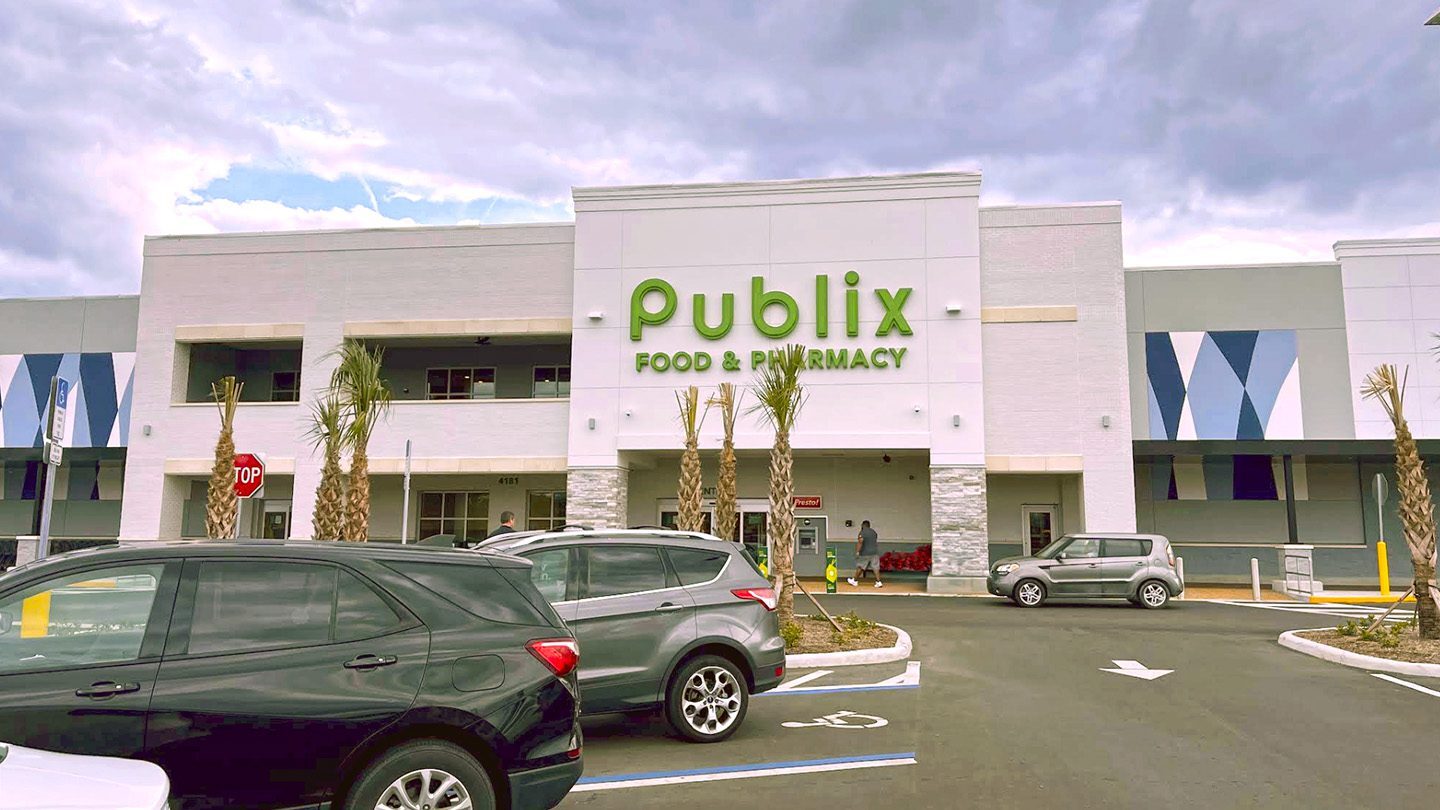 Exterior view of a Publix Food & Pharmacy store with a modern white facade, green signage, and palm trees planted along the front entrance. Several cars are parked in front, and a few customers are entering the store.