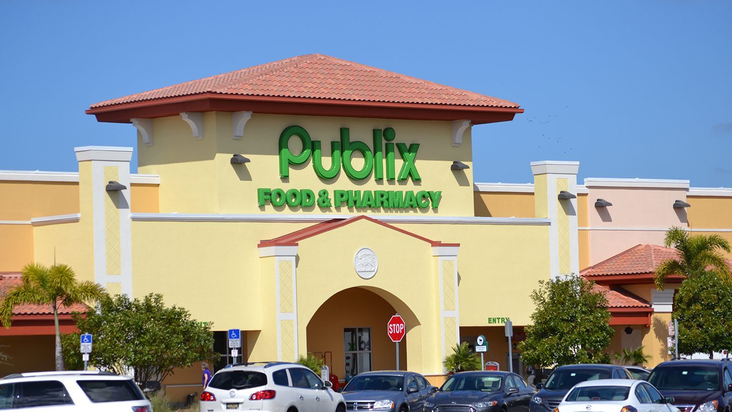 Frontal view of the Publix Food & Pharmacy entrance with bright yellow and cream exterior, red roof, arched entryway, and several cars parked outside.