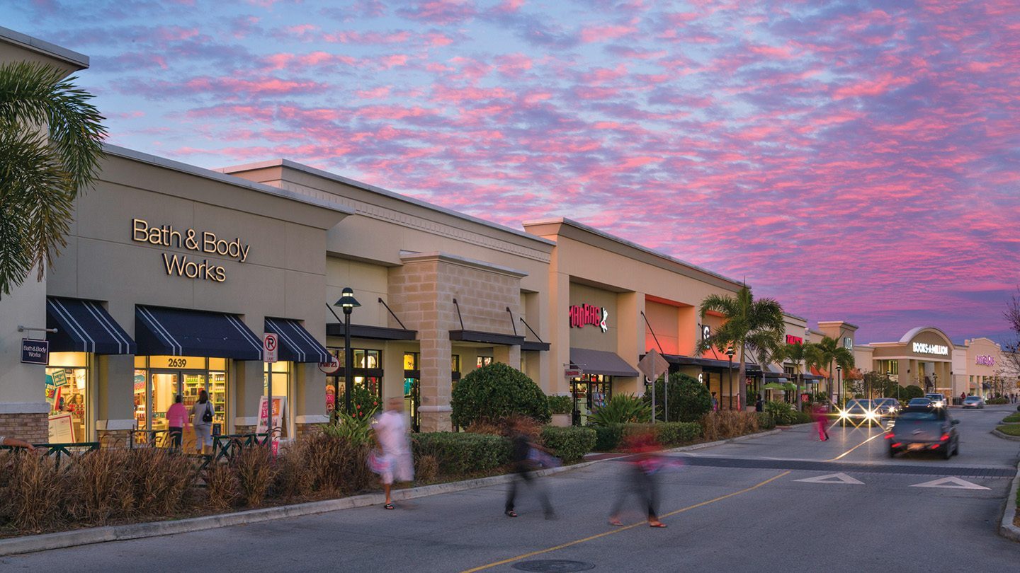 Evening view of a retail strip with a vibrant sunset sky painted in pink and purple hues. The storefronts include Bath & Body Works (with a visible address number "2639"), a store labeled "MADRAG" with a red neon sign, and Books-A-Million visible in the distance. Shoppers walk along the sidewalk and cross the street, some blurred in motion. Palm trees and decorative landscaping line the walkway, and cars are parked or driving past, with headlights on due to the low light.