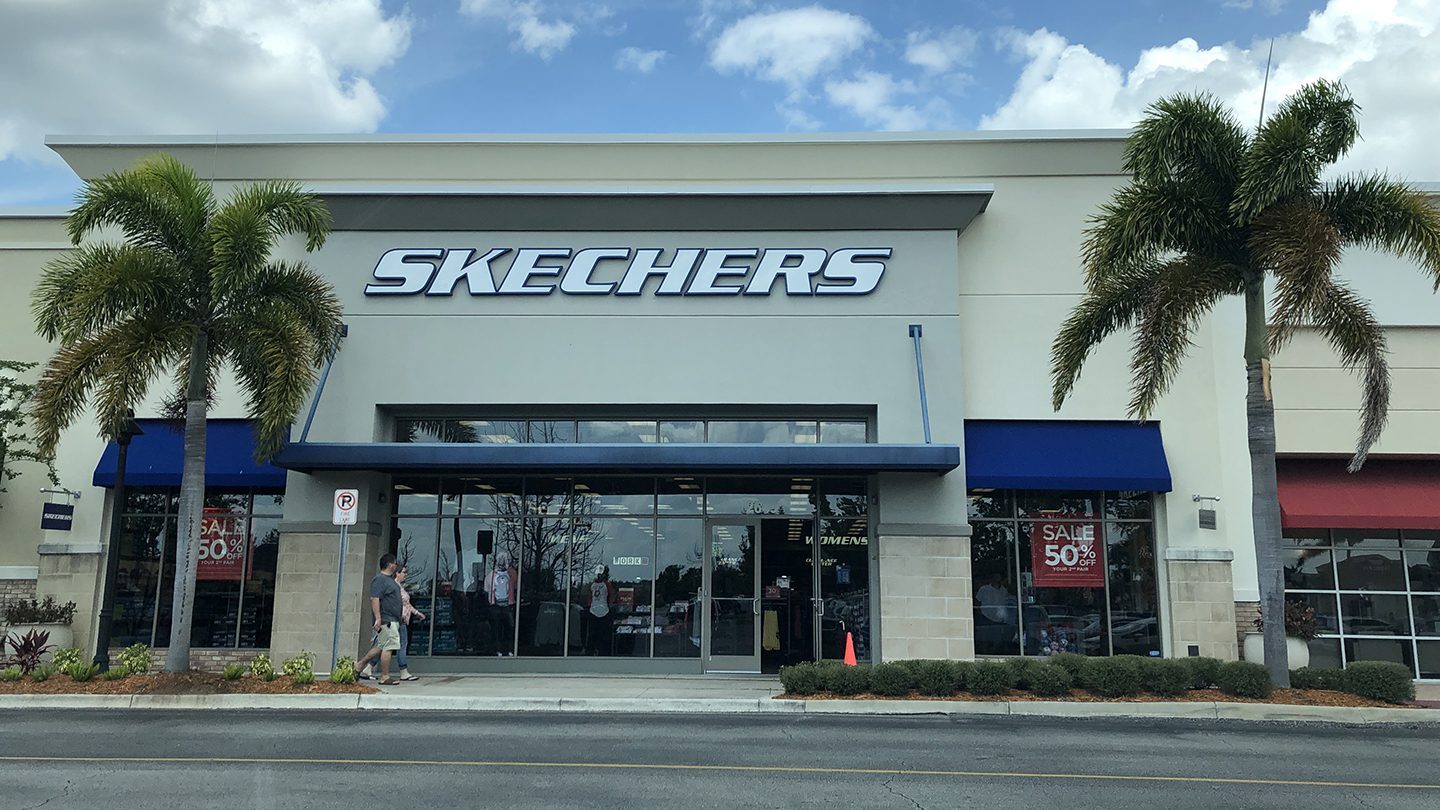 Front view of a Skechers retail store with large glass doors and windows, set between two palm trees under a bright, partly cloudy sky. The storefront features the brand name "SKECHERS" in large bold letters above the entrance, with a blue awning and two sale signs in the windows advertising "50% Off." A person is seen walking on the sidewalk in front of the store, and some clothing displays are visible inside.