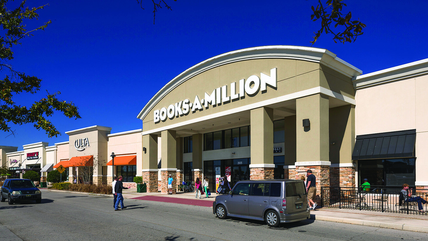 Street view of the Books-A-Million store with large white signage on a beige building featuring stone columns and a covered entrance. The store entrance has glass double doors and a small café sign reading "Joe Muggs Coffee" above the doorway. Promotional signs in the windows advertise sales, and a sidewalk with landscaping and a black streetlamp leads to the entrance. A black patio umbrella is visible on the right side of the building.