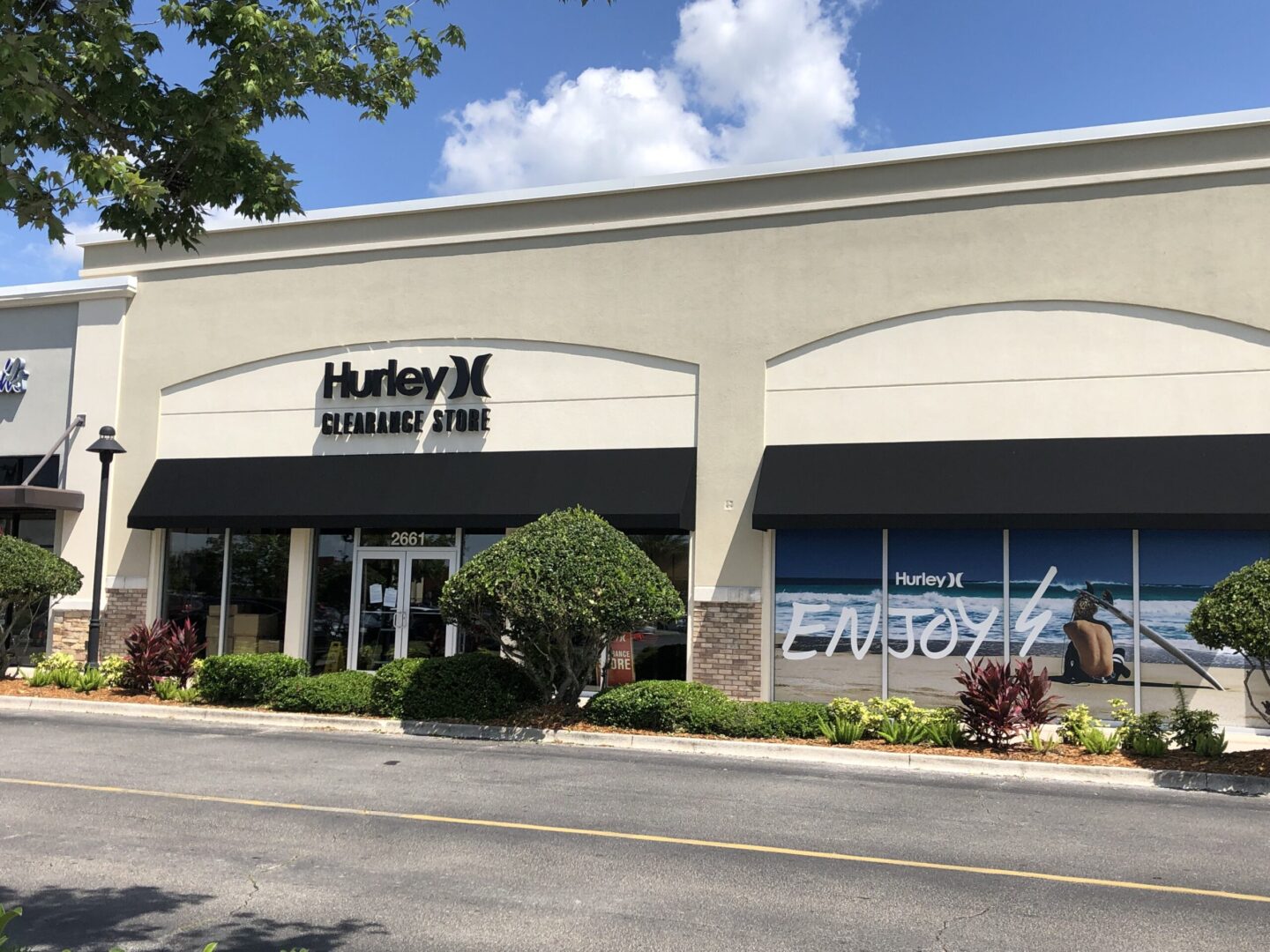 Front view of the Hurley Clearance Store featuring a light beige facade with black awnings over the windows and entrance. The store’s name and logo are prominently displayed above the door. A large window display on the right shows a photo of a surfer sitting on the sand with a surfboard, with the word “ENJOY” written in bold white letters across the beach scene. Well-trimmed bushes and landscaped greenery line the storefront, and a clear blue sky is visible above.Front view of the Hurley Clearance Store featuring a light beige facade with black awnings over the windows and entrance. The store’s name and logo are prominently displayed above the door. A large window display on the right shows a photo of a surfer sitting on the sand with a surfboard, with the word “ENJOY” written in bold white letters across the beach scene. Well-trimmed bushes and landscaped greenery line the storefront, and a clear blue sky is visible above.