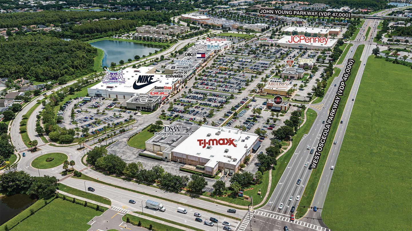 Aerial view of The Loop West shopping center in Kissimmee, Florida, showcasing a large retail plaza with major store signs including TJ Maxx, Nike, DSW, GameStop, Bath & Body Works, Burlington, and JCPenney. The shopping center is surrounded by expansive parking lots and lush greenery, bordered by West Osceola Parkway, a multi-lane highway with moderate traffic. Nearby, smaller restaurants such as Pei Wei and Abuelo’s are visible along with a central roundabout entrance and residential areas in the background.