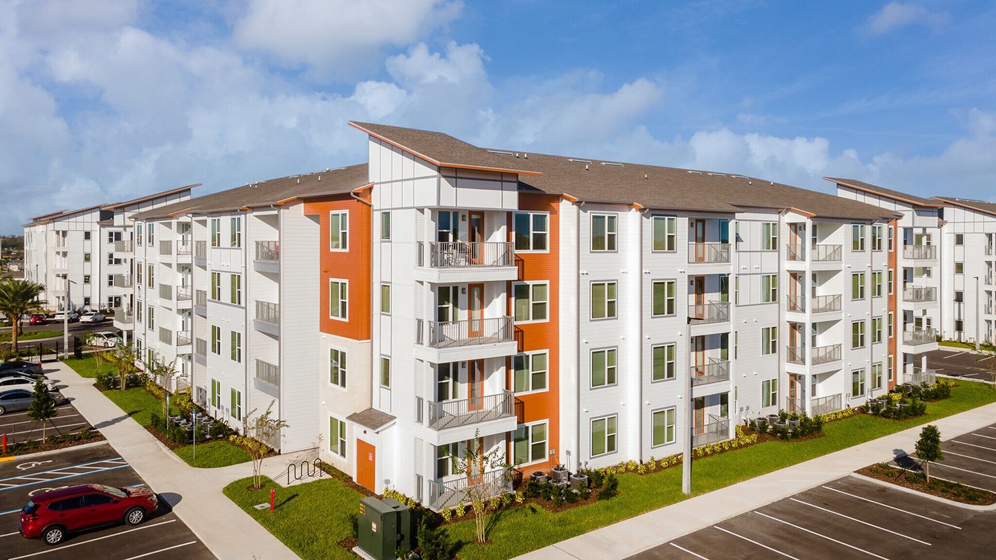A four-story modern apartment building with a flat roof and orange-and-white exterior panels. Each unit features large windows and private balconies. The building is surrounded by a neatly landscaped area with green grass, shrubs, and a sidewalk adjacent to a parking lot with marked spaces, including accessible parking.