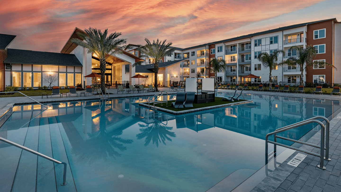 Ground-level view of a luxury apartment pool area at sunset, featuring a large pool with poolside lounge chairs, red umbrellas, and palm trees. A clubhouse with large glass windows and a modern four-story apartment building are illuminated in the background.