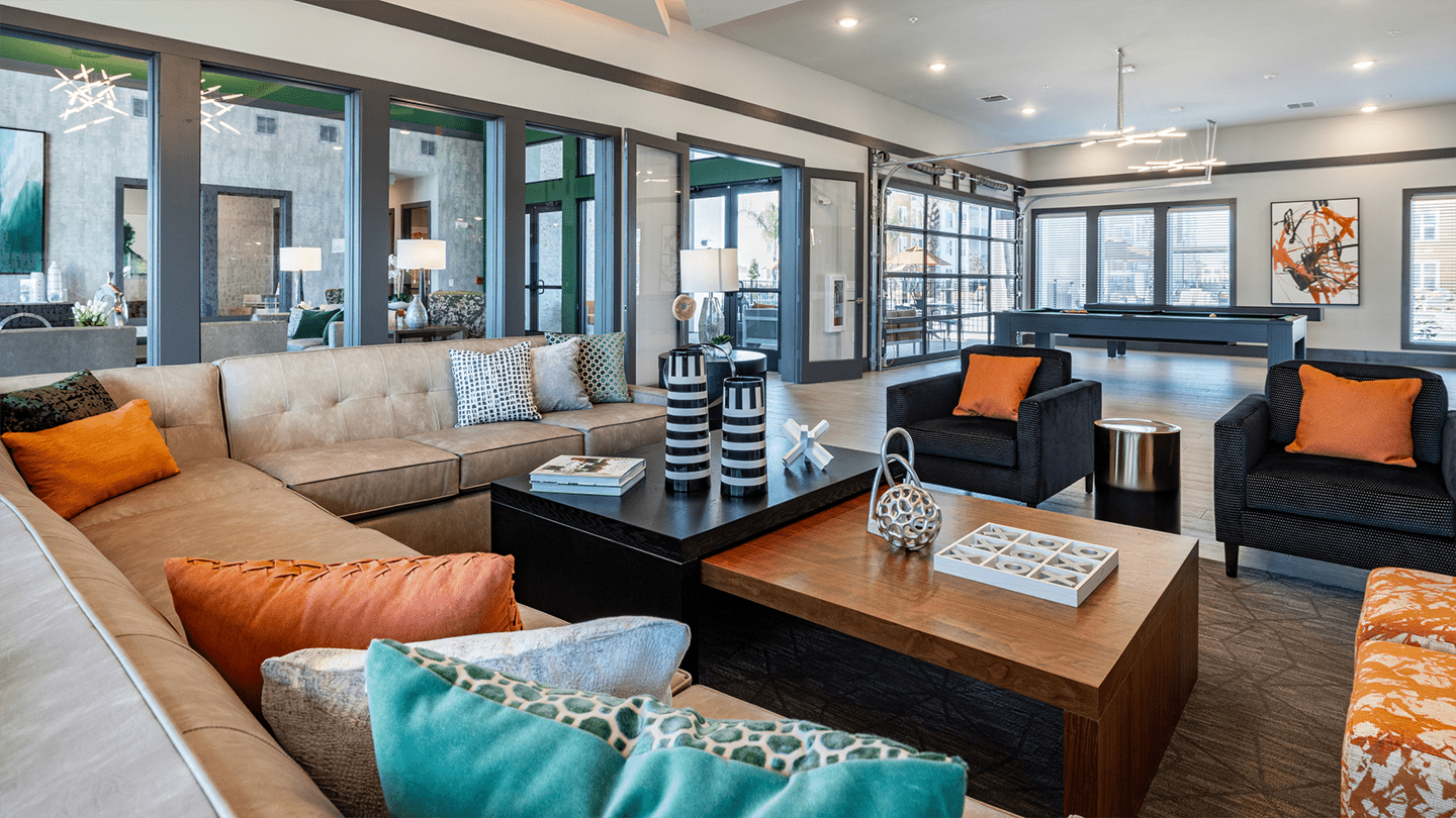 Modern apartment community lounge featuring a large beige sectional sofa with colorful throw pillows, a wooden coffee table with decorative accents, and a pool table in the background. The room has large windows and glass doors allowing natural light to fill the space.