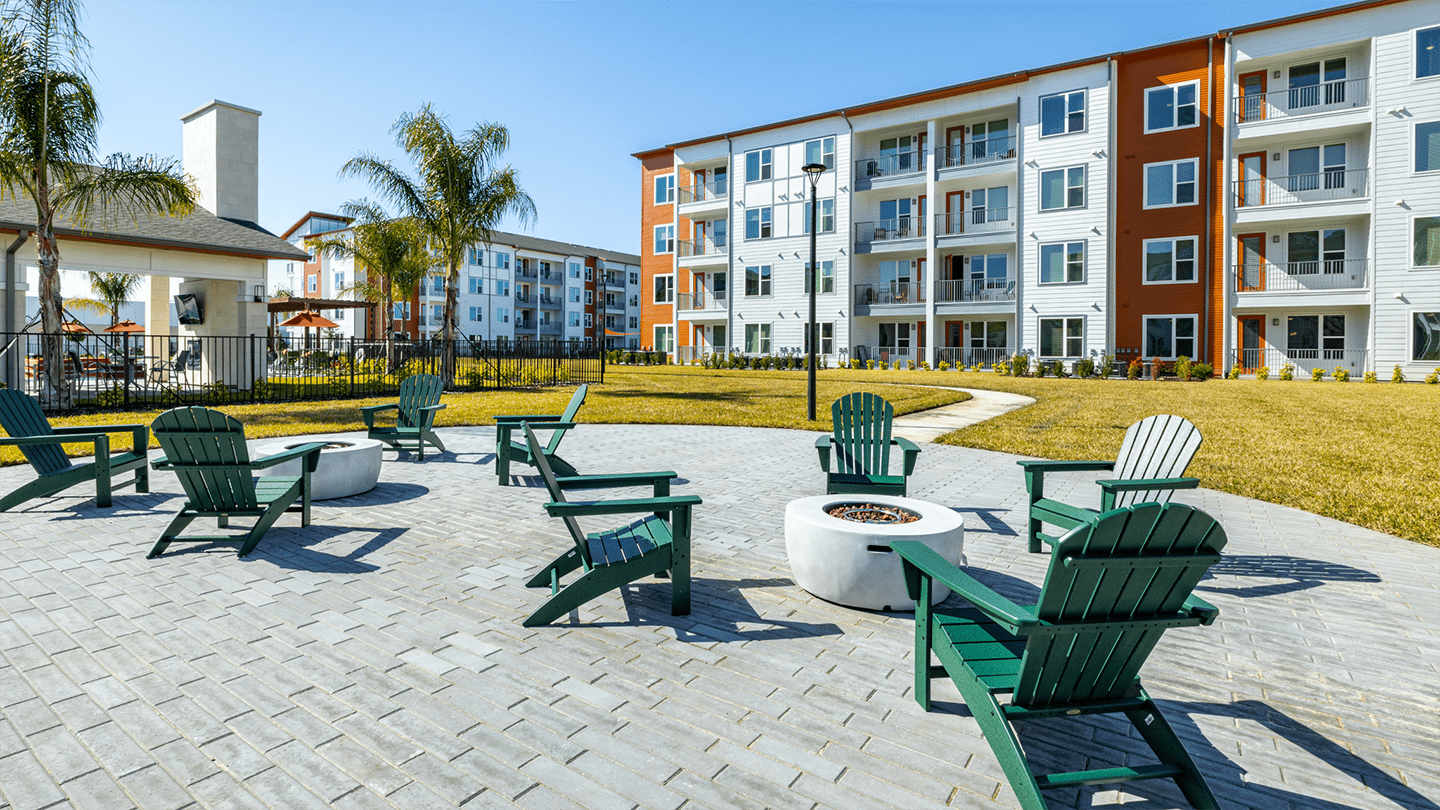Outdoor seating area with green Adirondack chairs arranged around circular white fire pits on a paved patio, surrounded by well-maintained lawns and palm trees. Modern four-story apartment buildings with balconies are visible in the background under a clear blue sky.